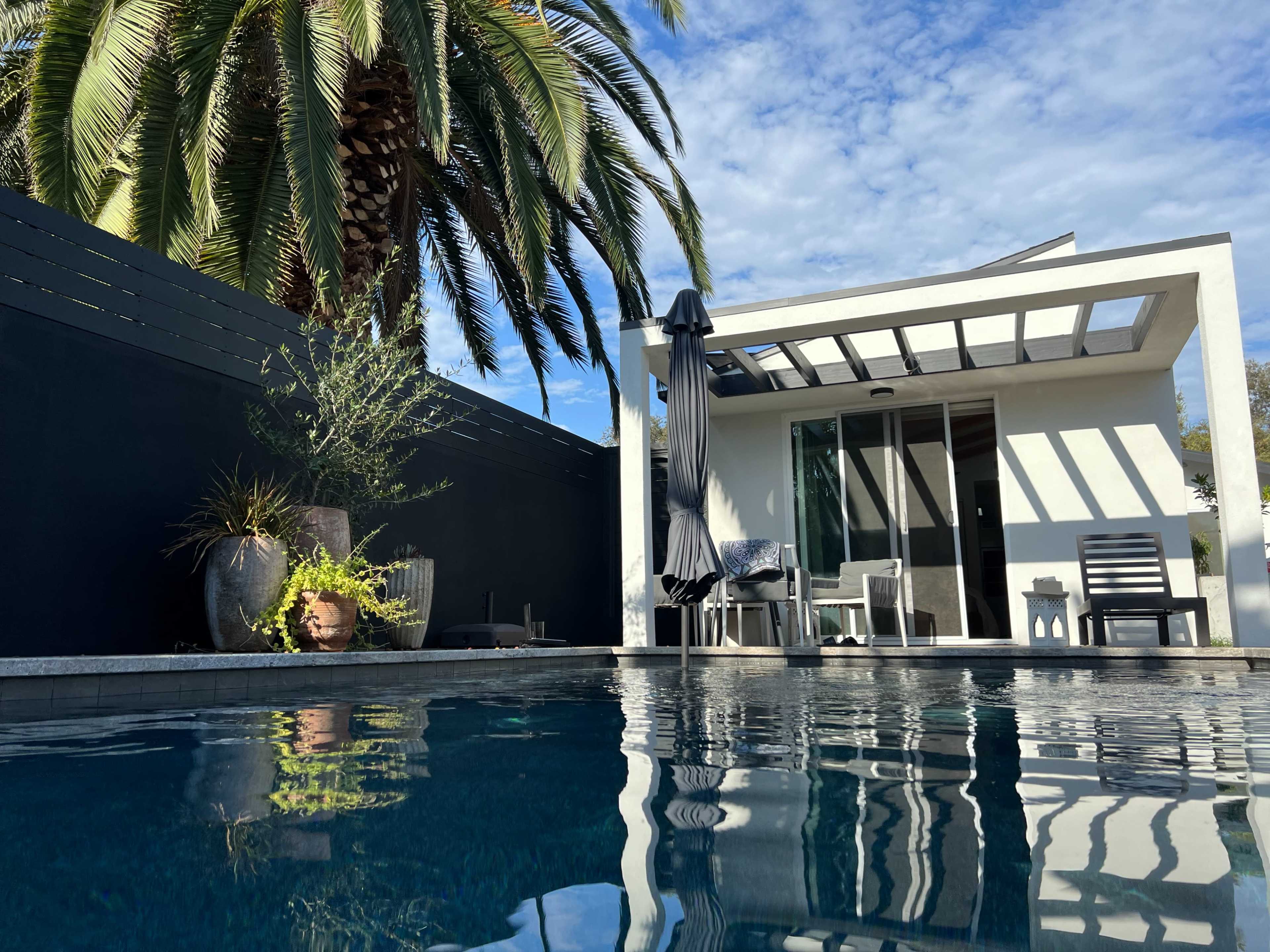 A modern poolside area featuring a minimalist structure, palm trees, and patio furniture.