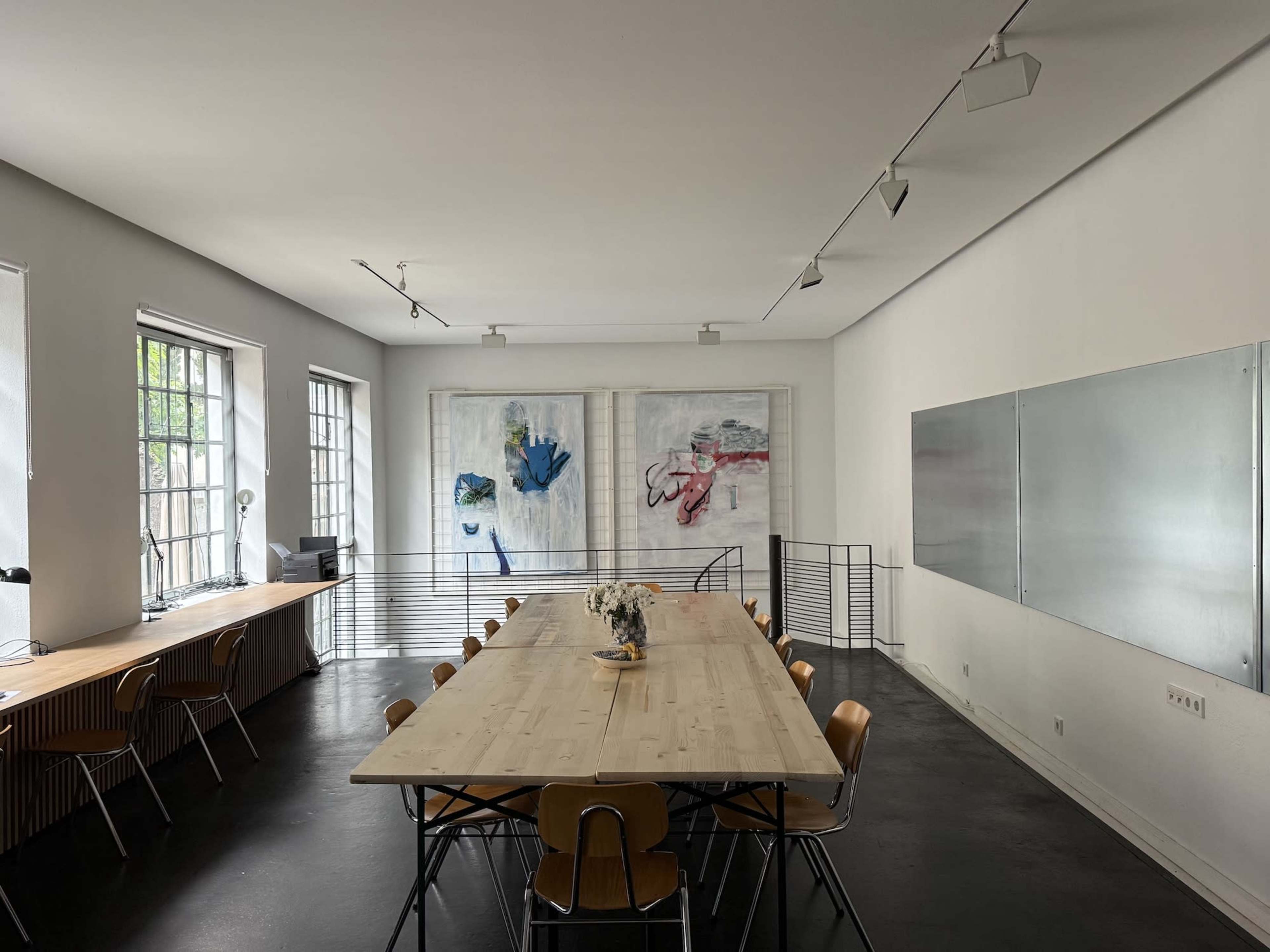 A spacious office meeting room features a long wooden table surrounded by wooden chairs, two large abstract paintings on the wall, and a whiteboard on the adjacent wall.