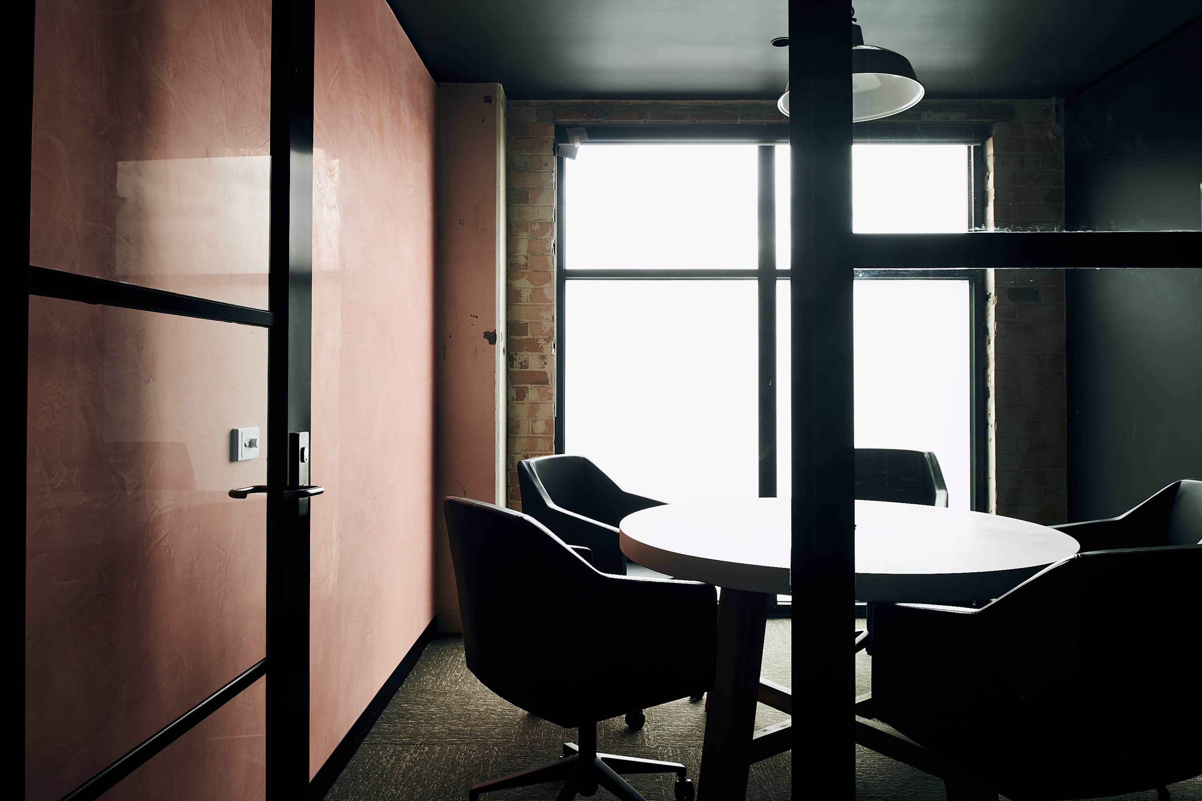 A conference room features a round table surrounded by black chairs, illuminated by natural light filtering through large windows.