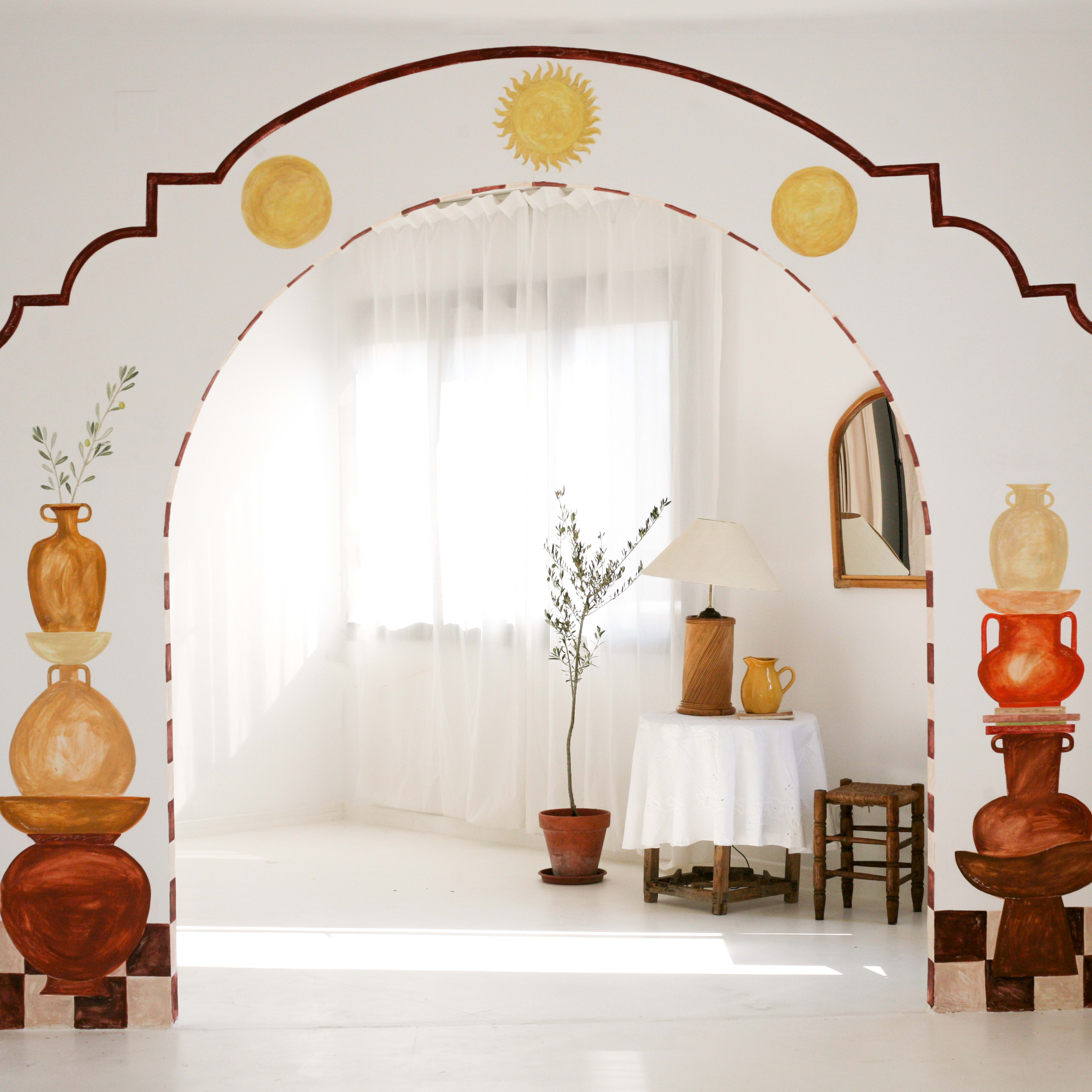 A white room features an archway decorated with painted pottery and a sun motif, leading to a space with a table, lamp, and potted plant.