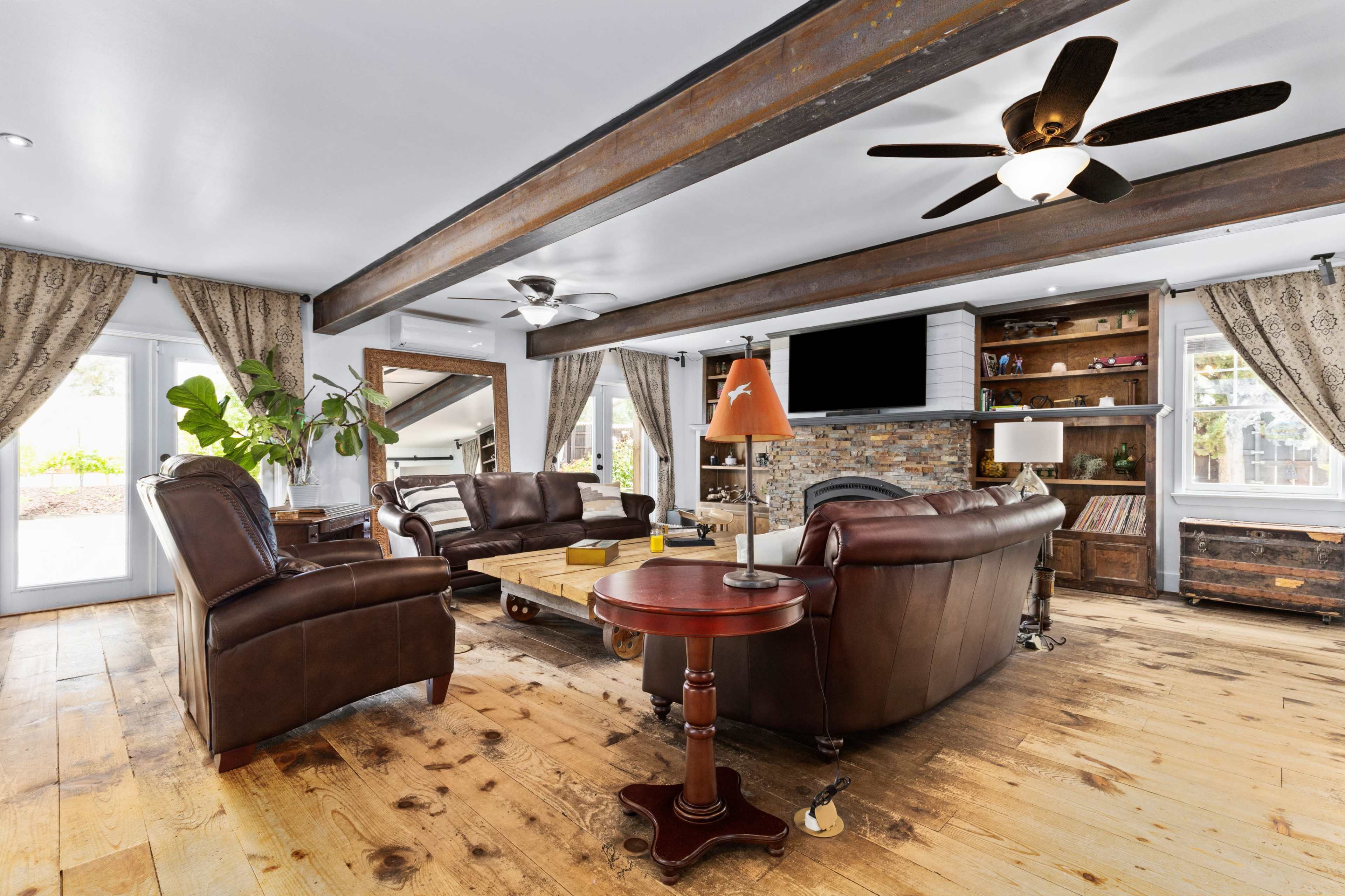 The living room features two leather sofas, a coffee table, a side table, and a stone fireplace, with wood beams overhead and large windows letting in natural light.