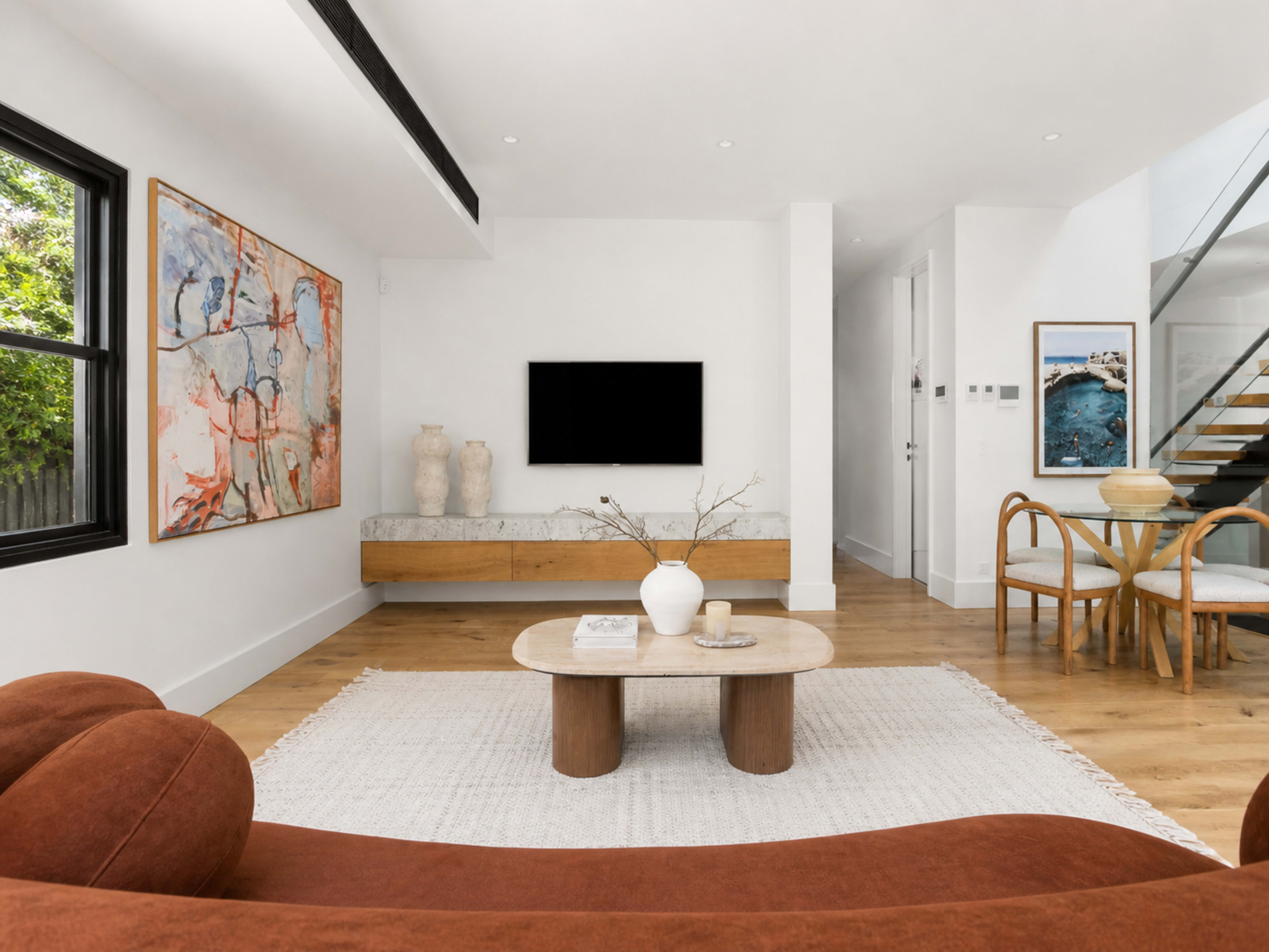 The living room features a large artwork on the wall, a modern coffee table, and a minimalist media console beneath a wall-mounted TV, with wooden flooring and a staircase visible in the background.