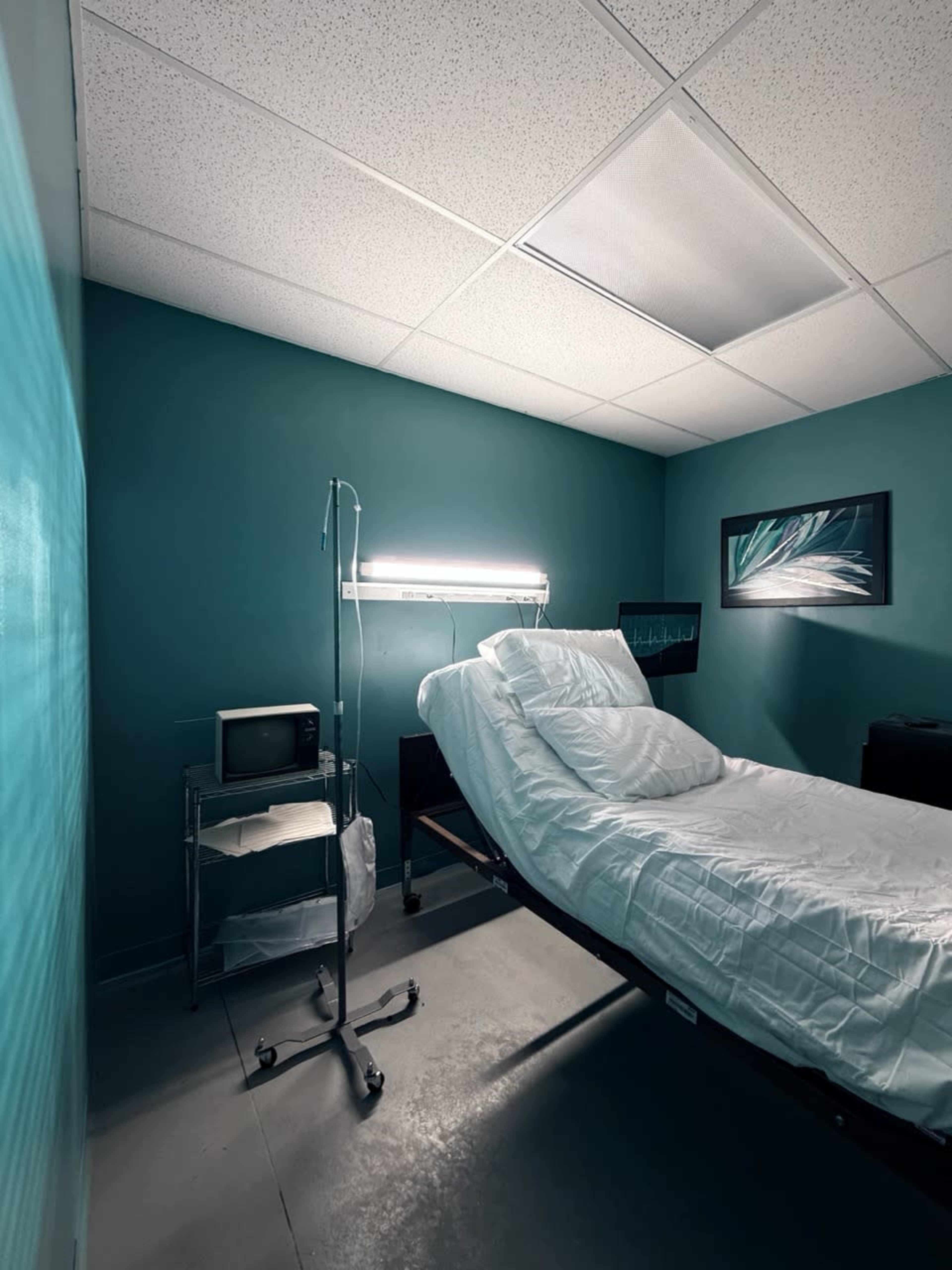 The image shows a hospital room with a single bed, a small vintage monitor on a cart, and teal-colored walls.