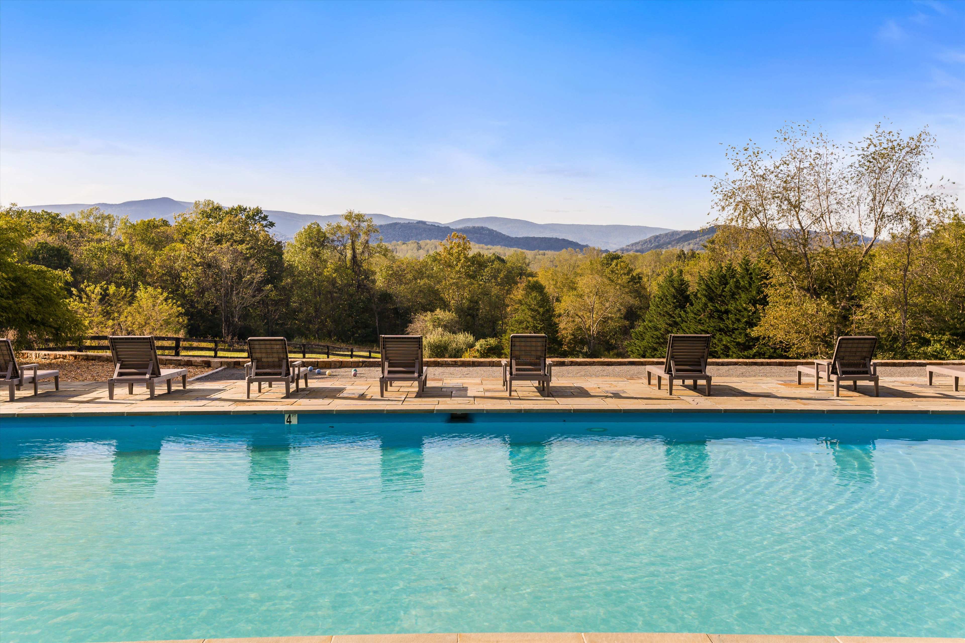 The image depicts a tranquil swimming pool surrounded by lounge chairs, with a scenic view of mountains and trees in the background.