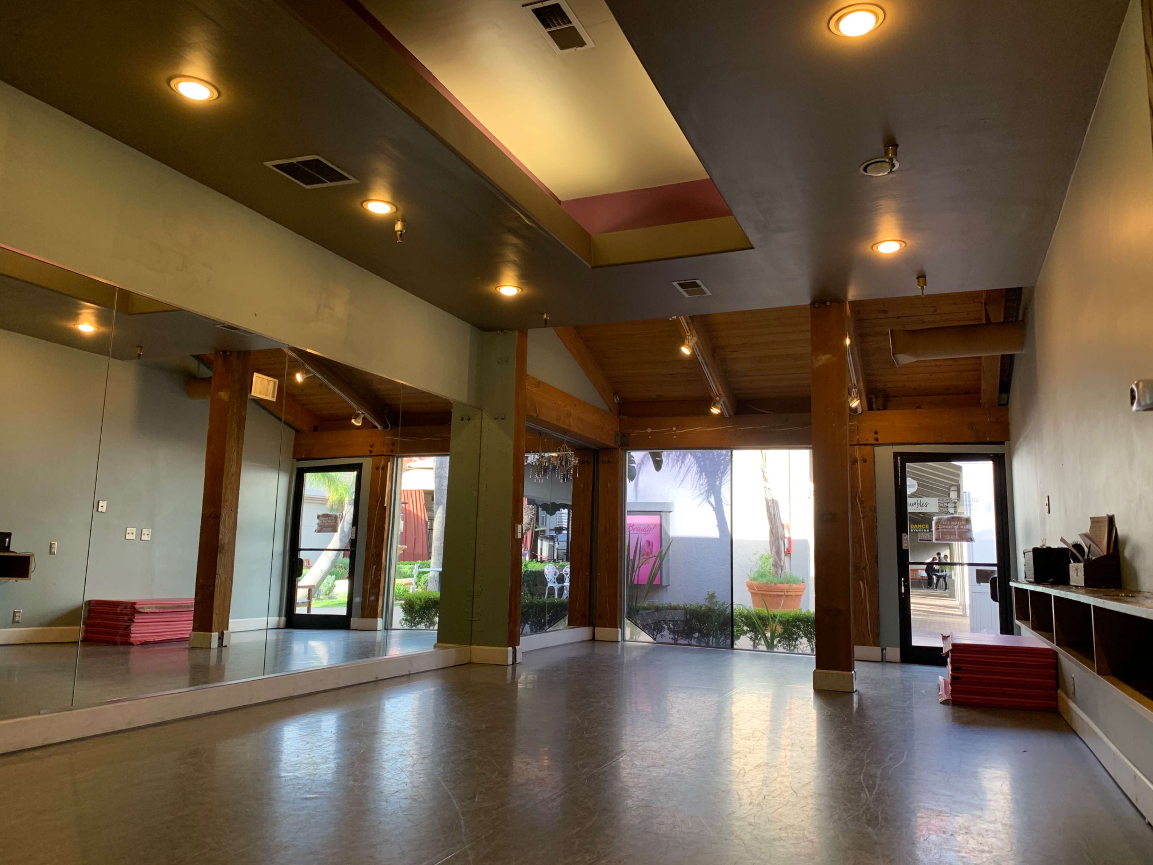 An empty dance studio features mirrored walls, wooden beams, and a large window overlooking an outdoor space.