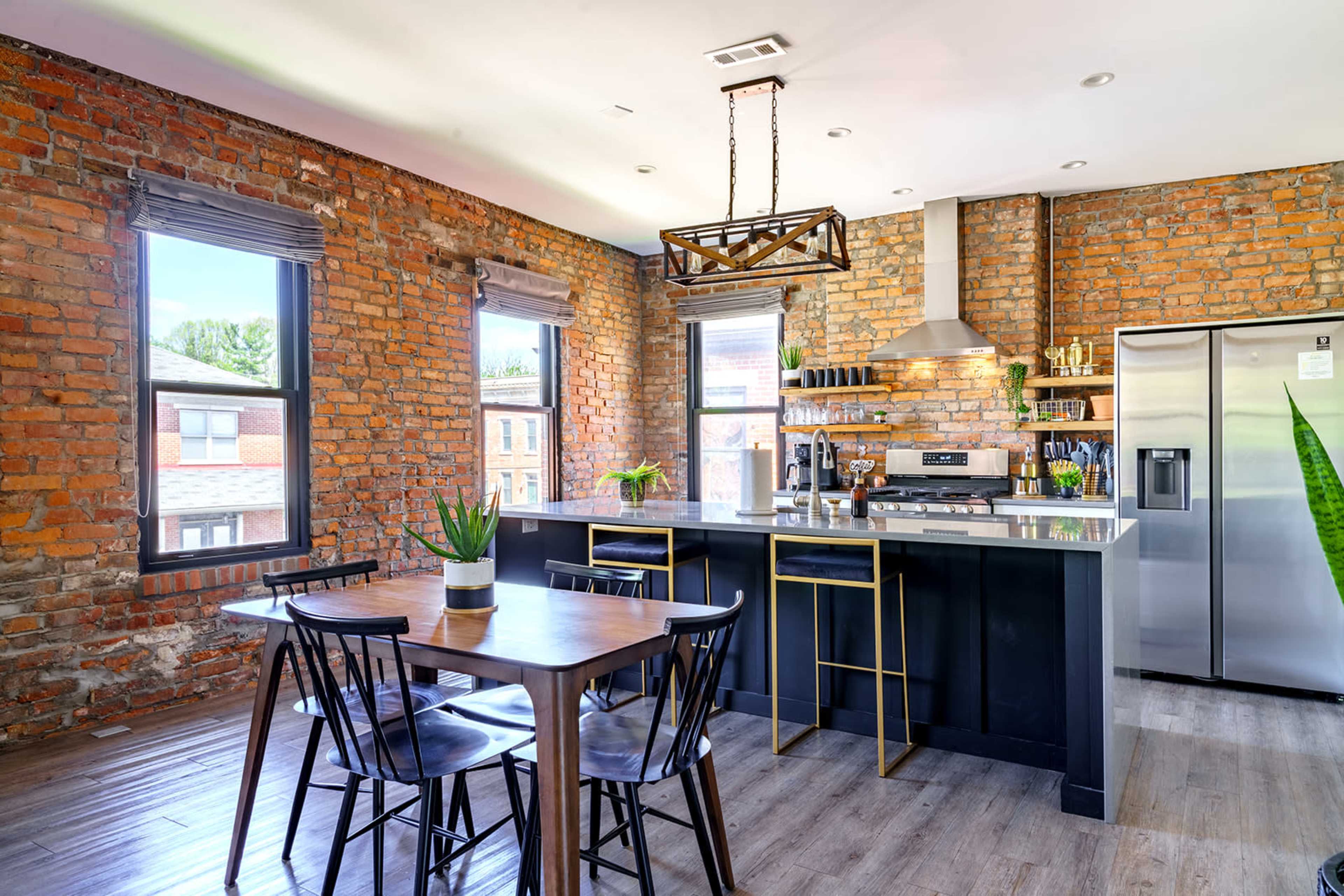 The image shows a modern kitchen with exposed brick walls, a central island, stainless steel appliances, and a wooden dining table with black chairs.