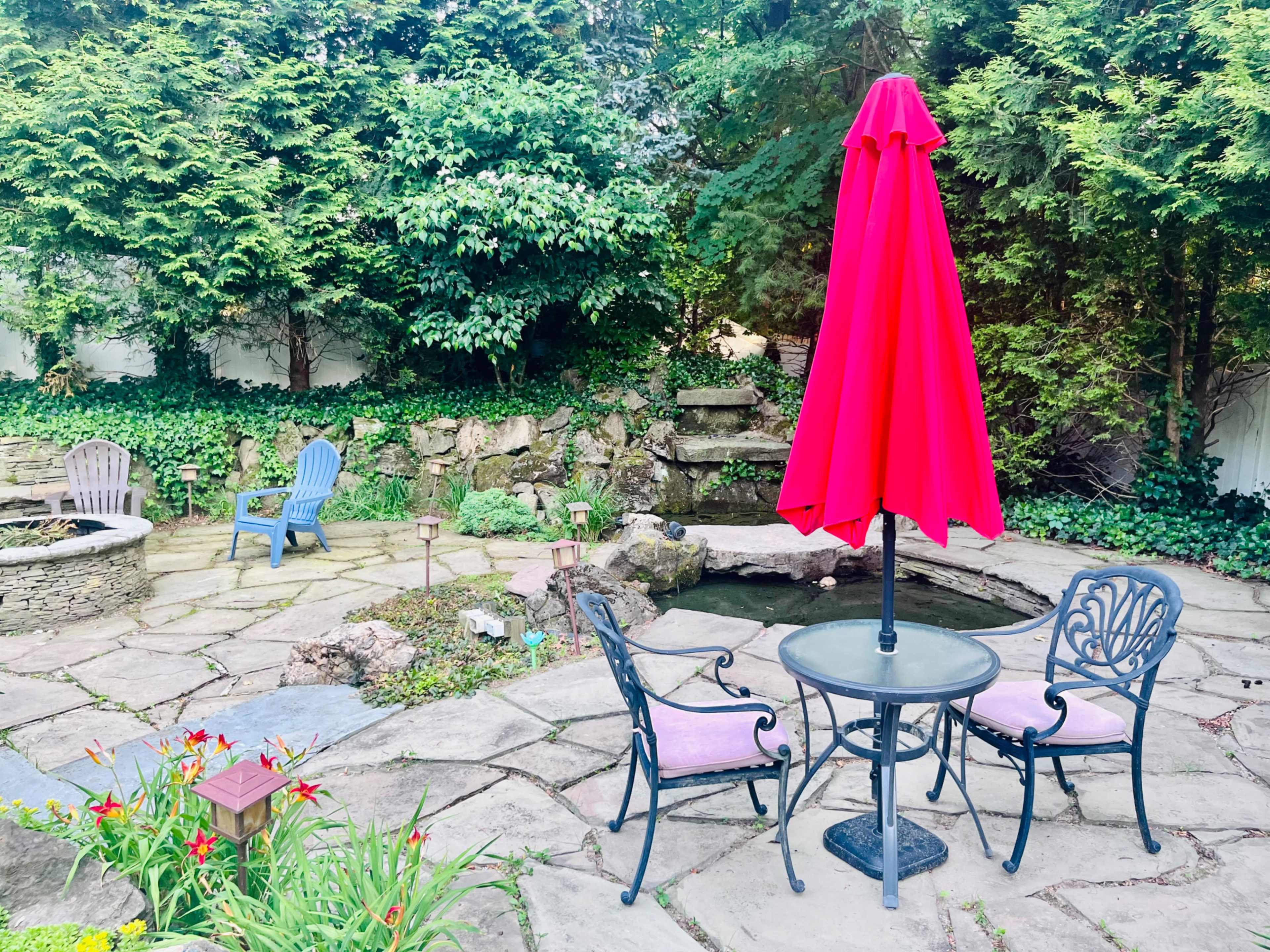 A circular table with two chairs and a red umbrella stands on a stone patio near a pond and landscaped greenery.