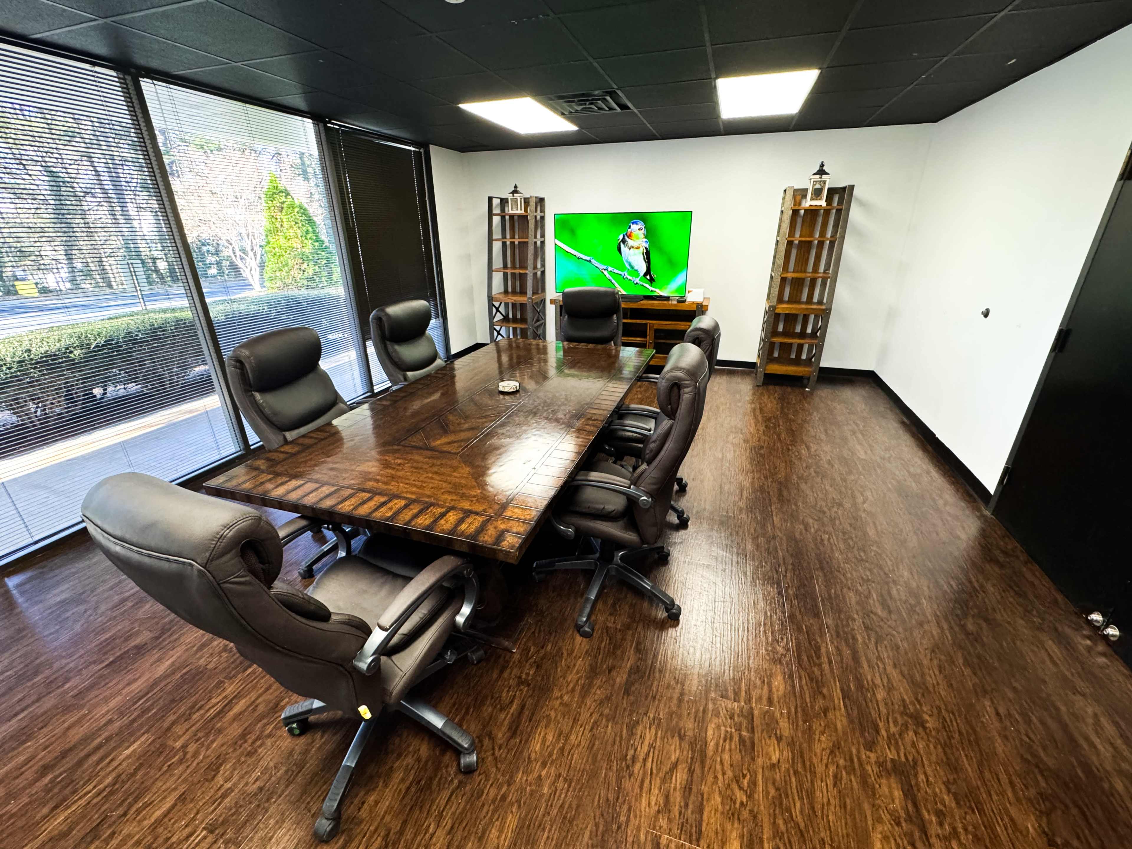 A conference room features a large wooden table surrounded by six ergonomic chairs, with a television and bookshelves in the background.