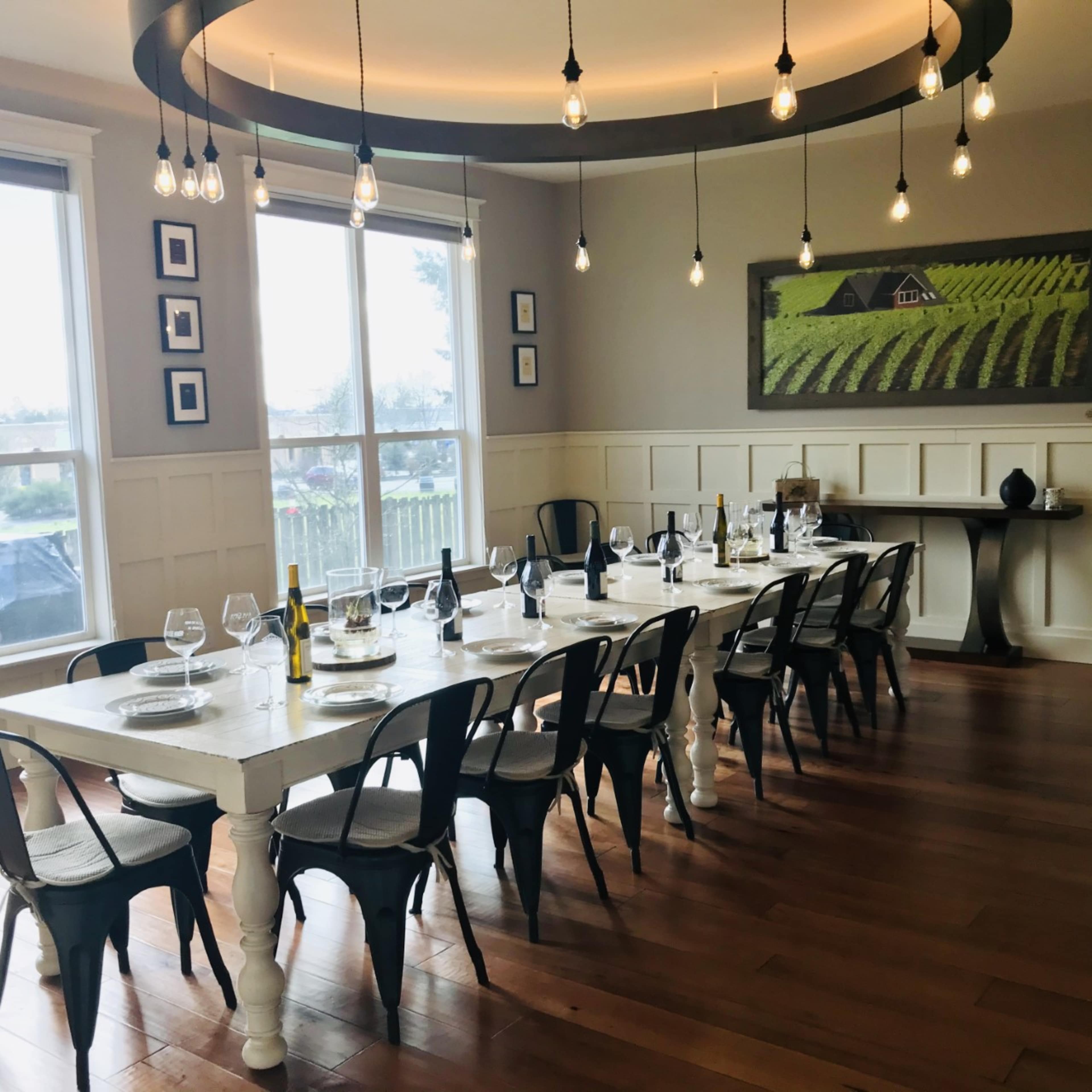 A long dining table is set with wine bottles and glasses in a well-lit room featuring large windows and artwork of a vineyard on the wall.