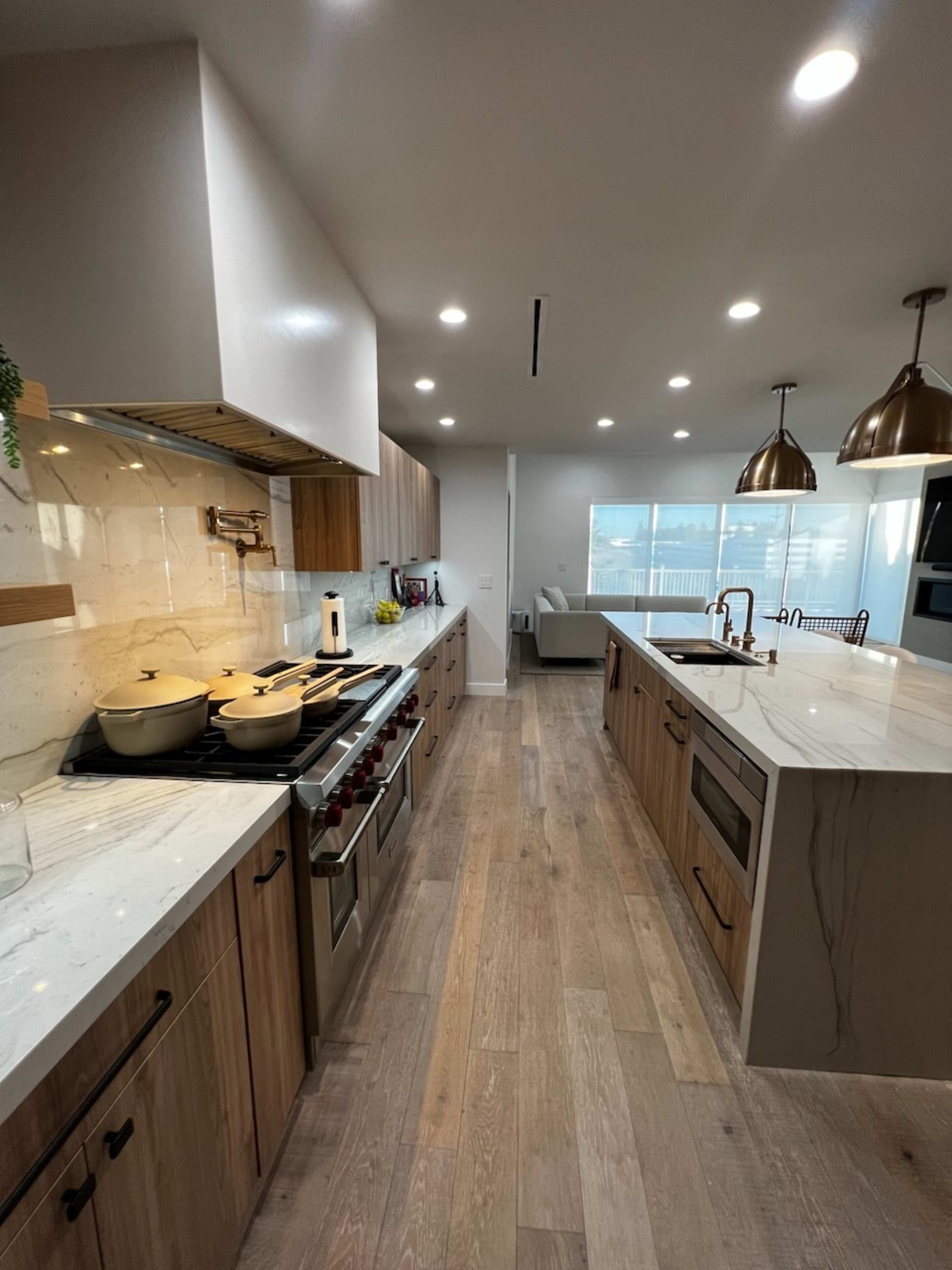 A modern kitchen with wooden cabinets, a marble countertop, and multiple cooking appliances, leading to a bright living area with large windows.
