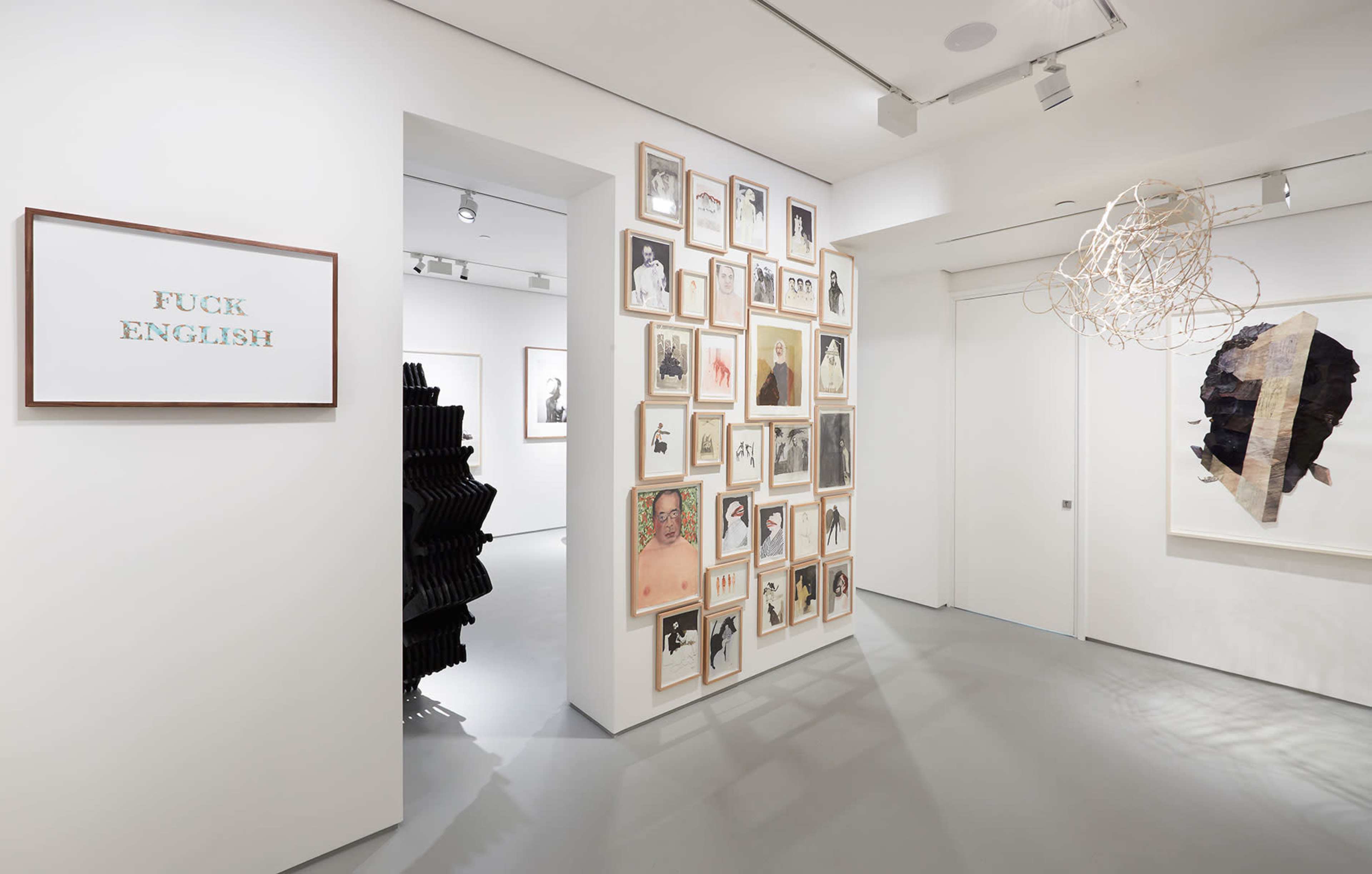 The image shows a modern art gallery featuring framed photographs and mixed media pieces on white walls, with a sign that reads "FUCK ENGLISH" displayed prominently.