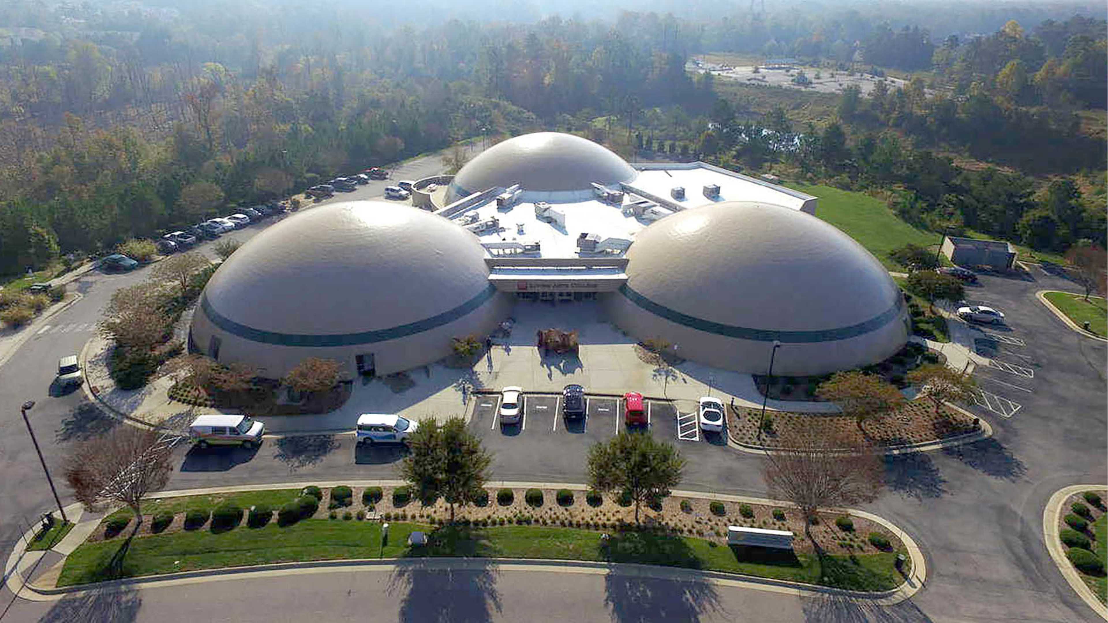 The image shows a building with three dome-shaped roofs surrounded by a parking lot and landscaped greenery.
