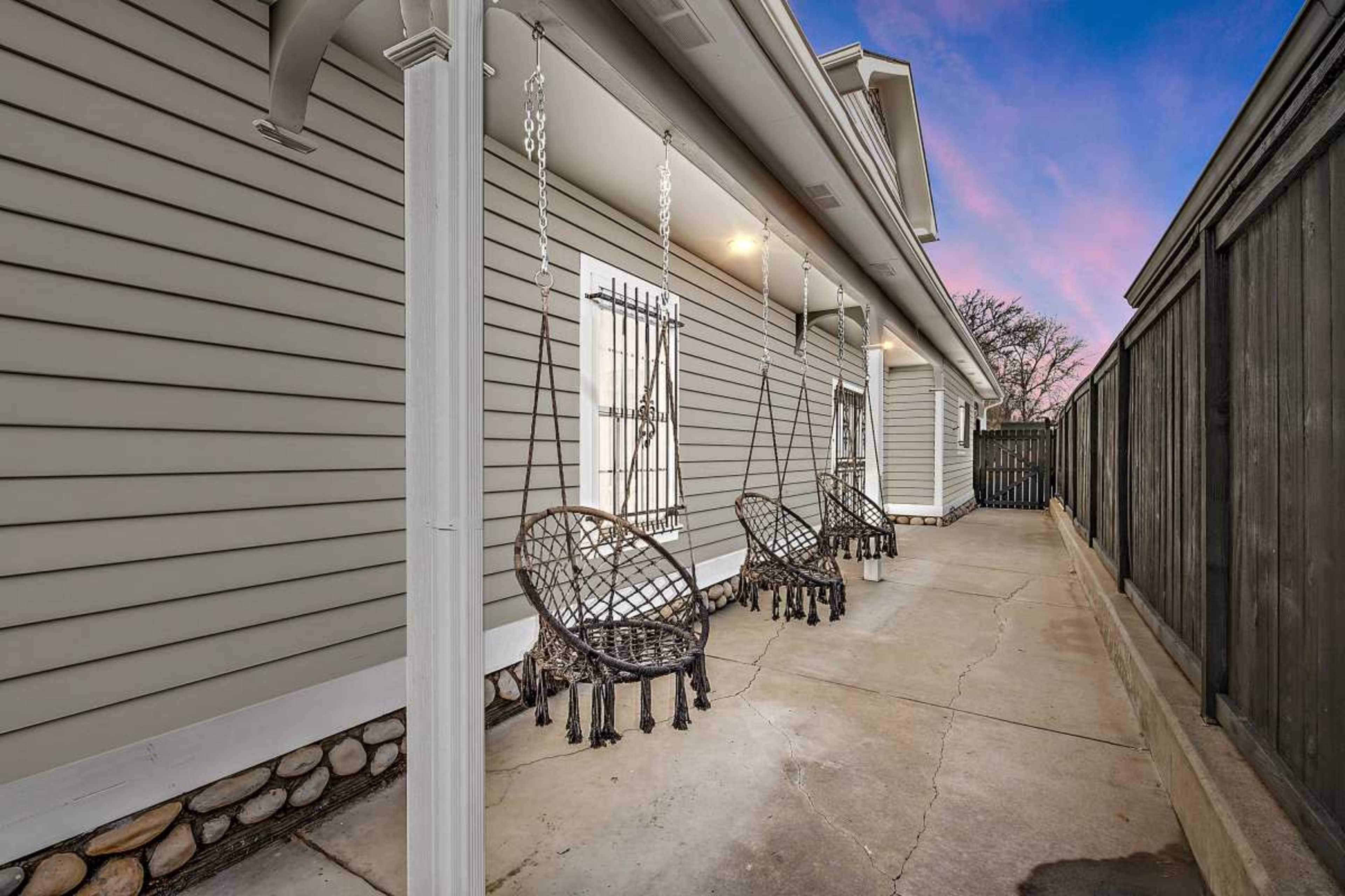 A side exterior of a house featuring three hanging chairs on a concrete walkway beside a wooden fence.