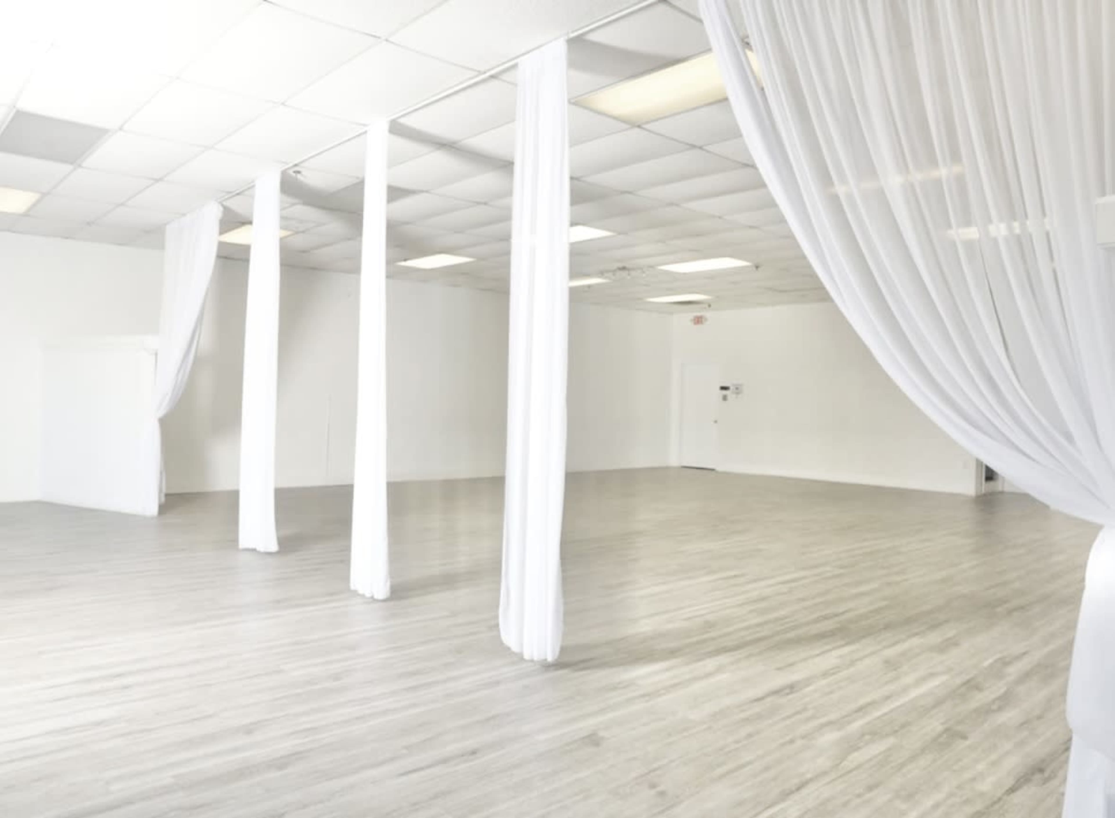 The image shows a large, empty studio space with white walls, a wooden floor, and draped white fabric hanging from the ceiling.