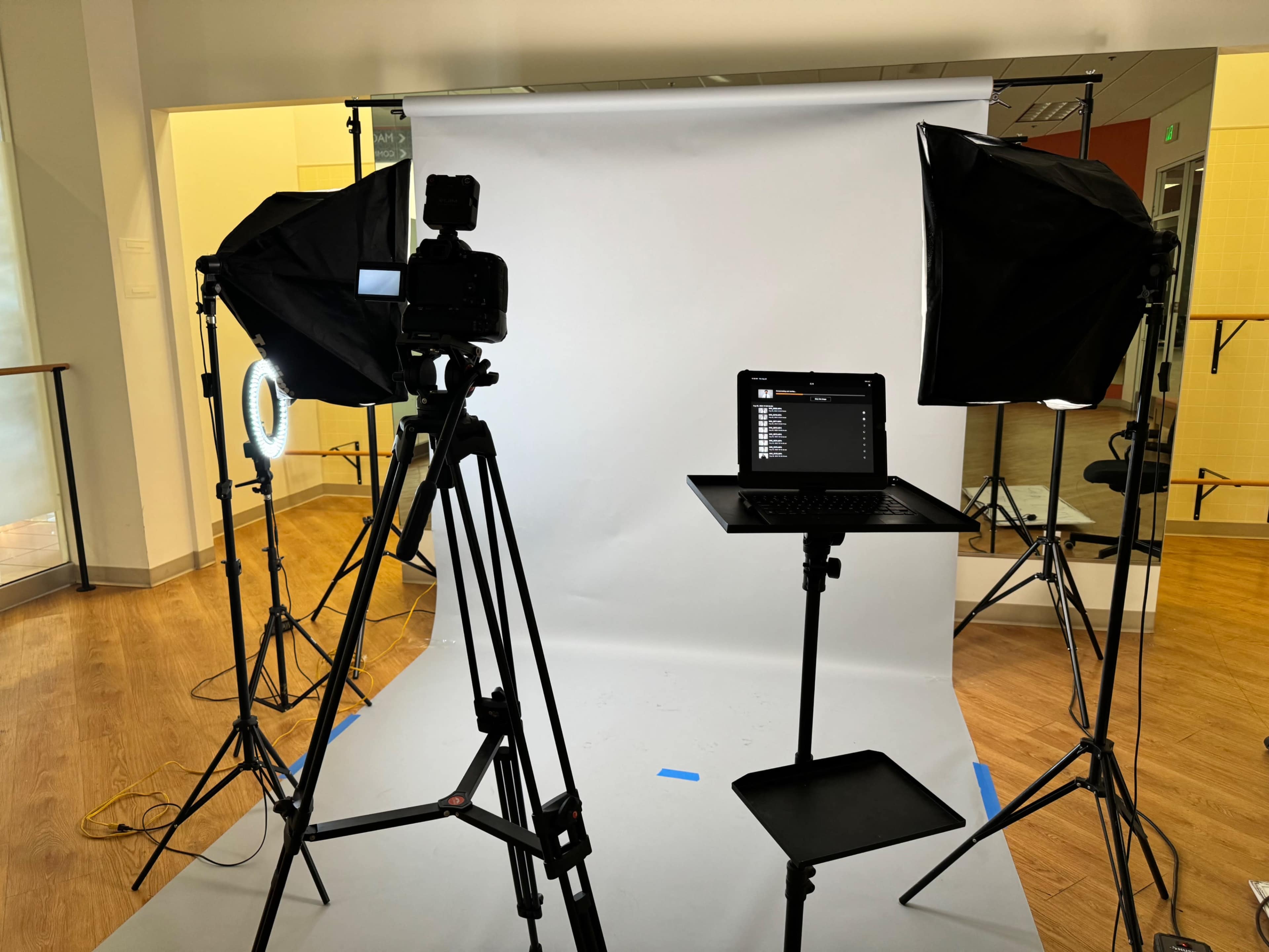 The image shows a photography setup with a camera on a tripod, two softbox lights, and a laptop on a stand in front of a plain white backdrop.