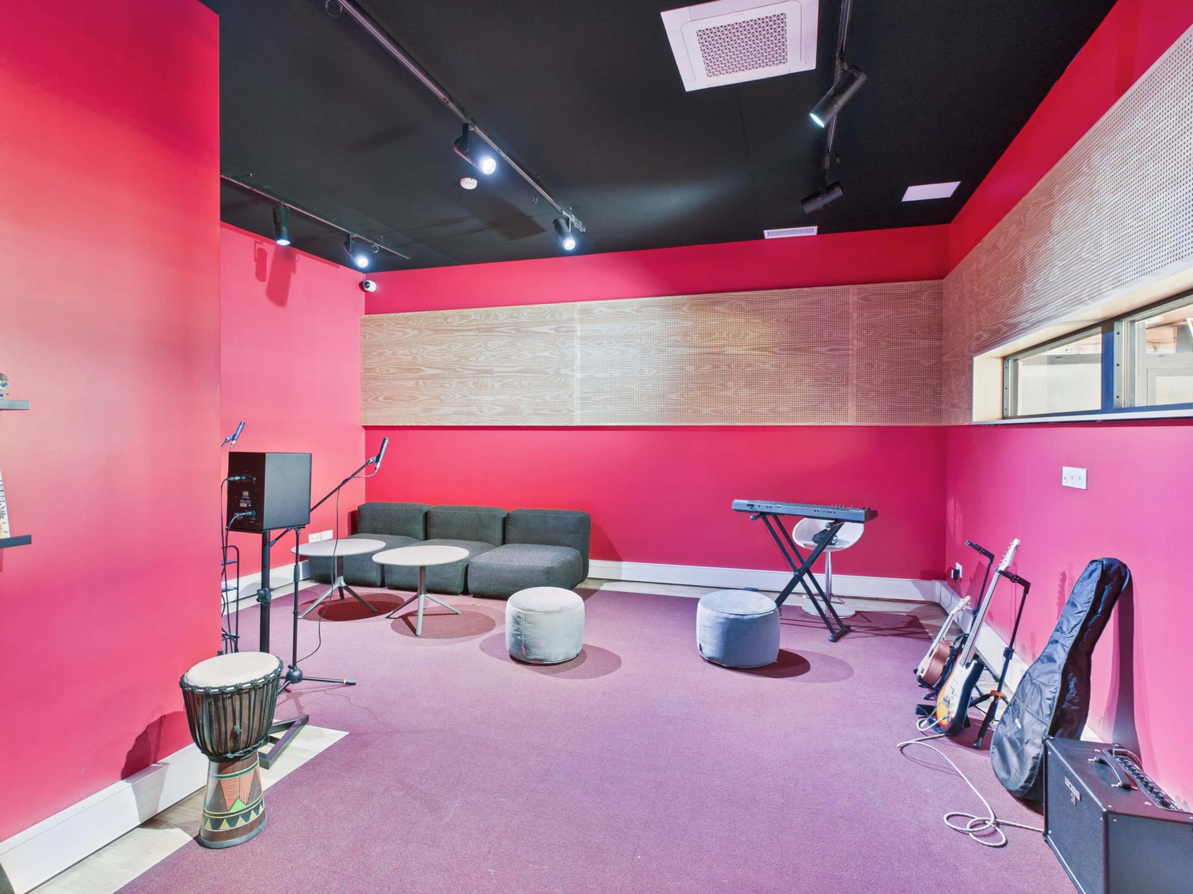 A small rehearsal room features red walls, a couch, a keyboard, and various musical instruments.