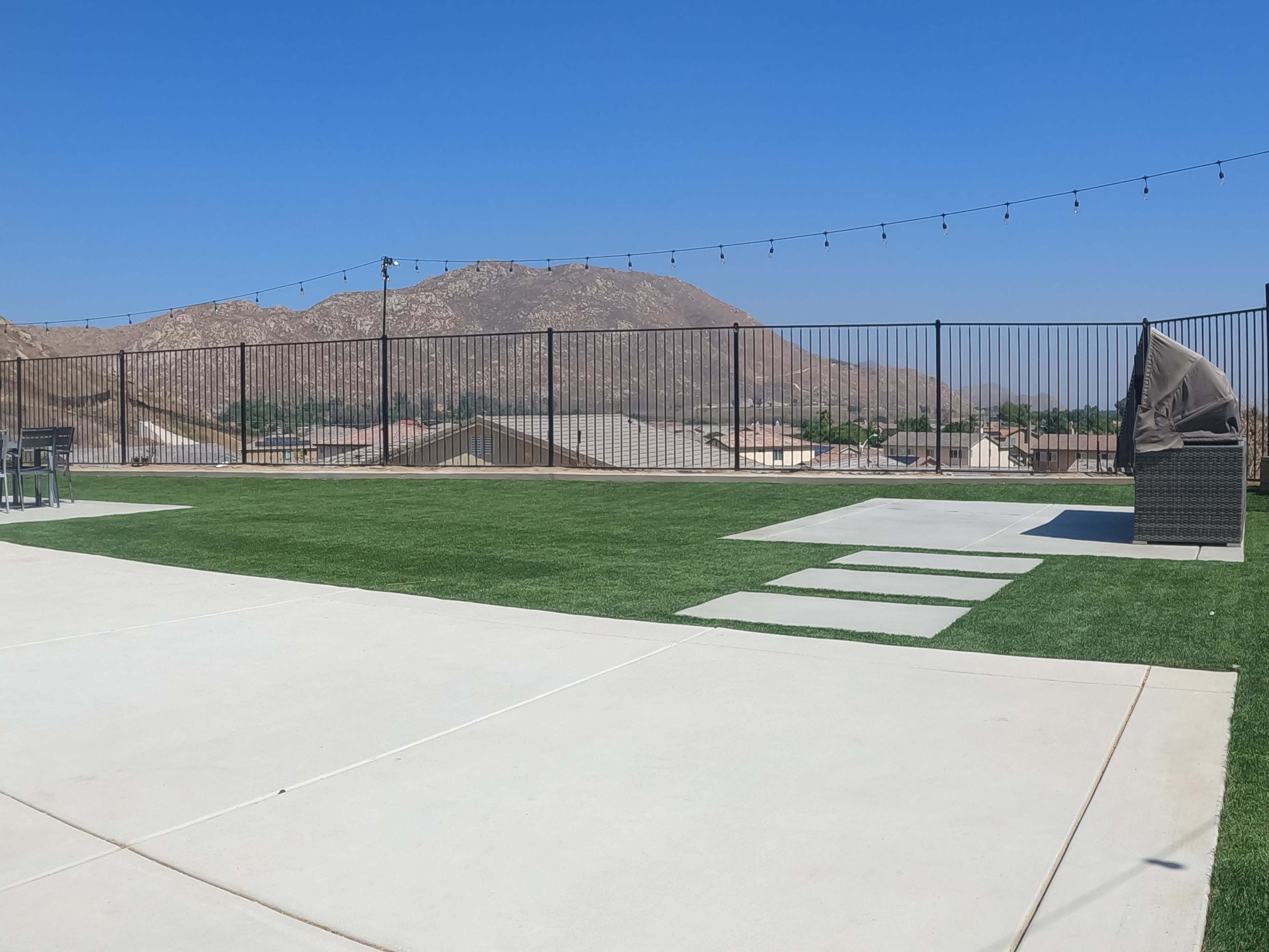 The image shows a fenced outdoor area with artificial grass, concrete pathways, and a mountain backdrop under a clear blue sky.