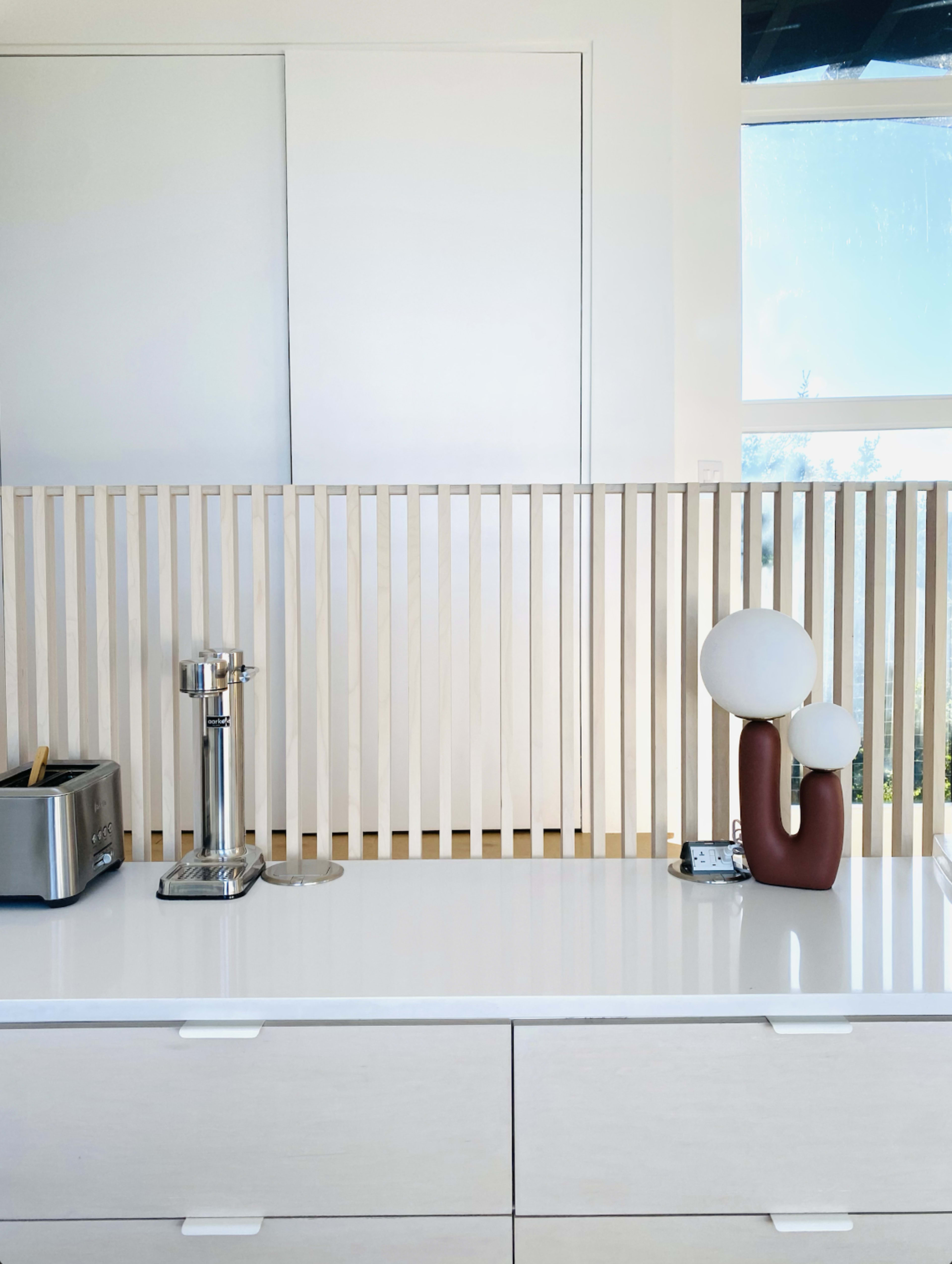 A sleek kitchen countertop features a toaster, a beverage tap, and a decorative lamp behind a wooden slatted partition.