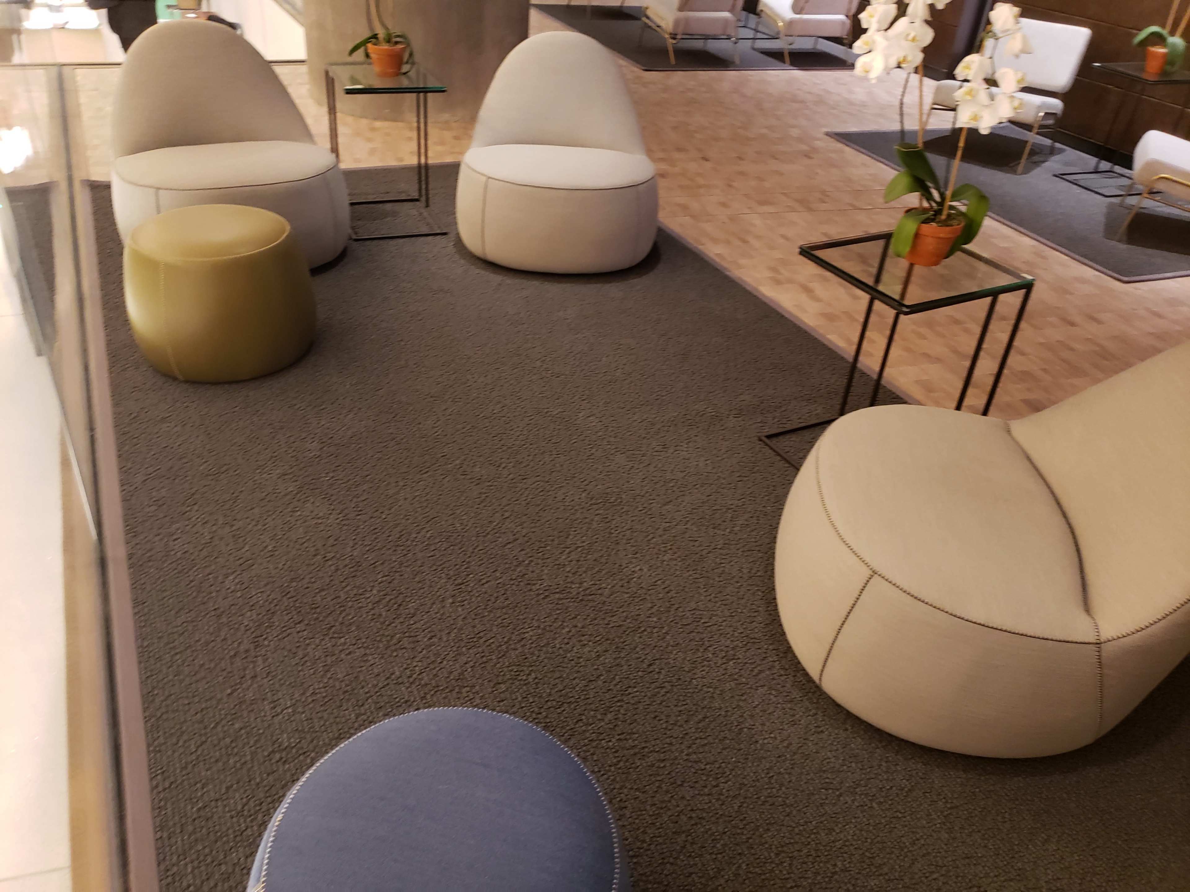 The image shows a modern lounge area featuring three light-colored seating pods, a small round gold stool, and two glass side tables on a gray carpet.
