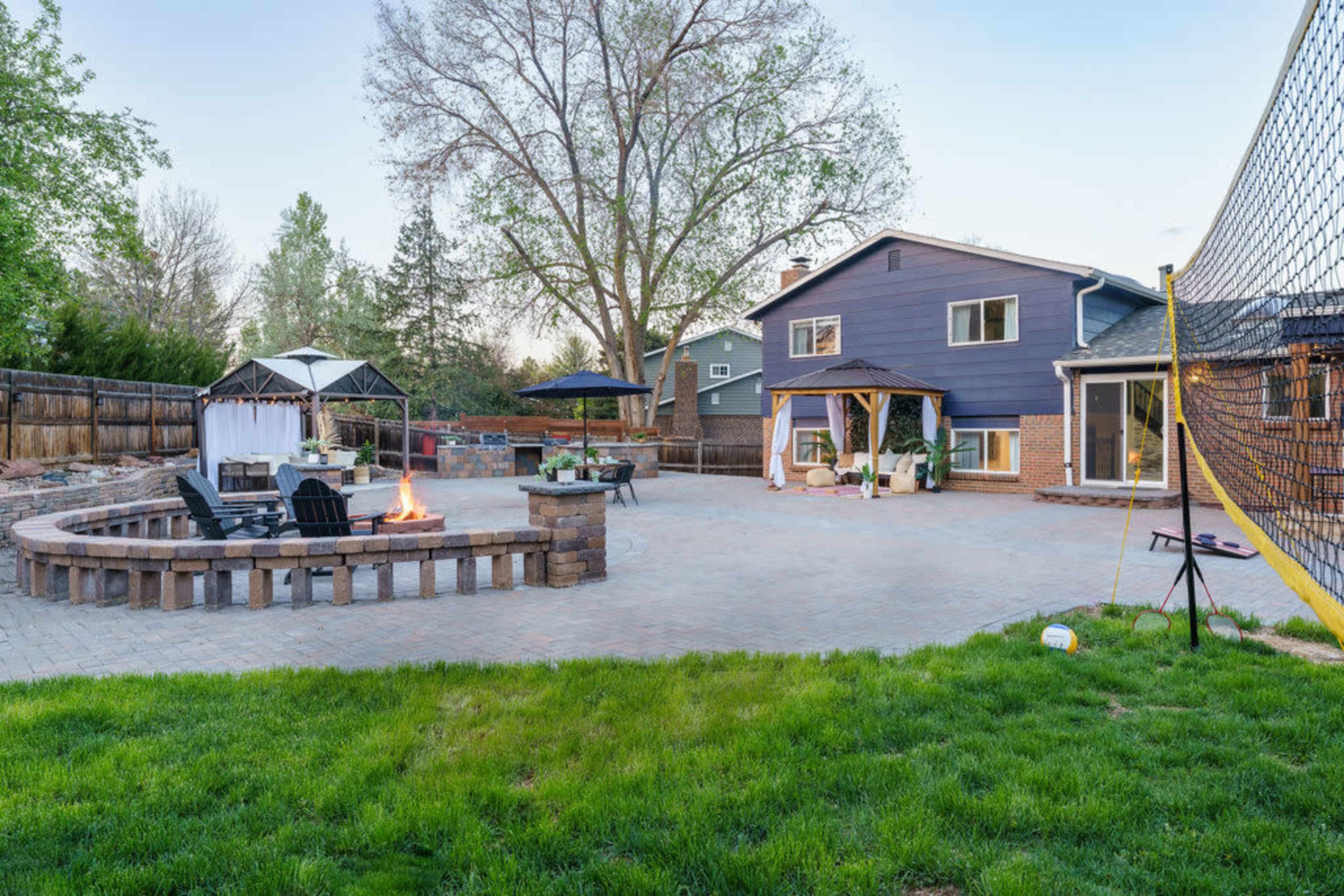 The image shows a backyard patio area with a round stone firepit surrounded by chairs, a gazebo, and a two-story house in the background.
