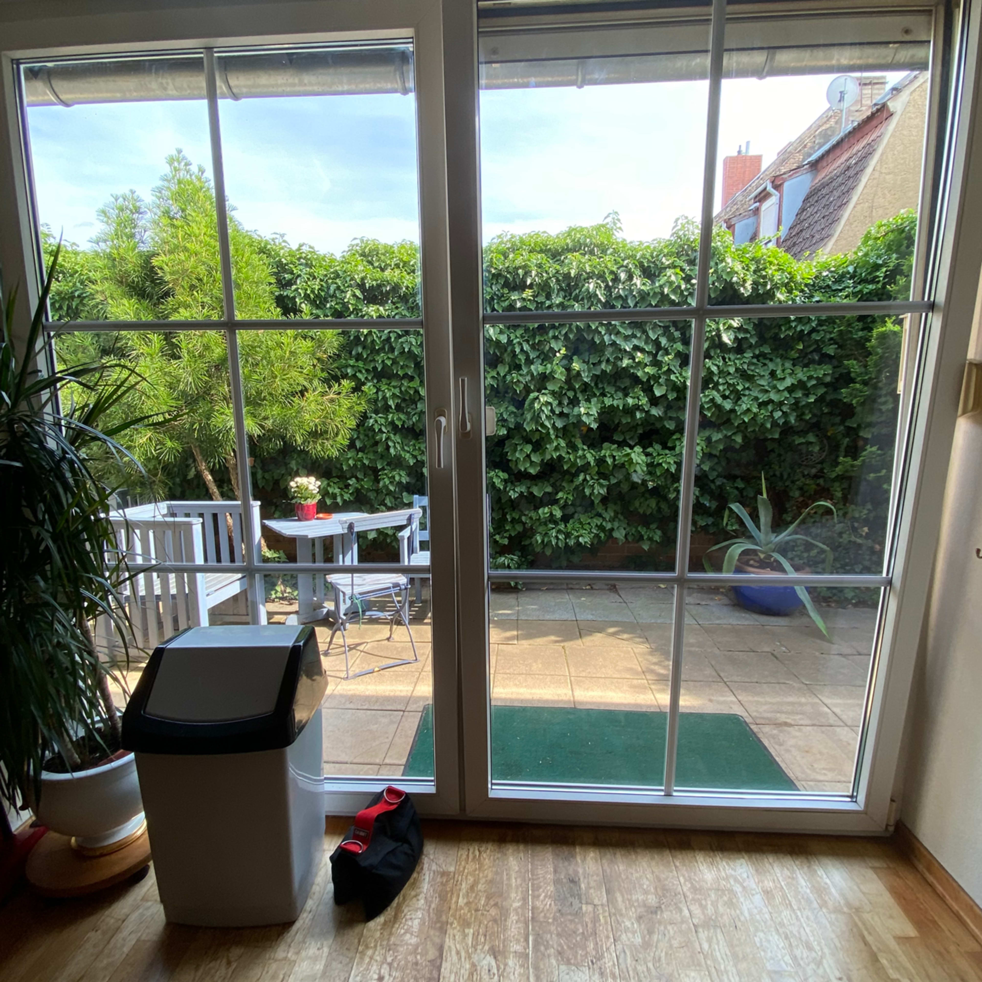 The image shows a view through a sliding glass door onto a green garden with outdoor furniture and a small table.
