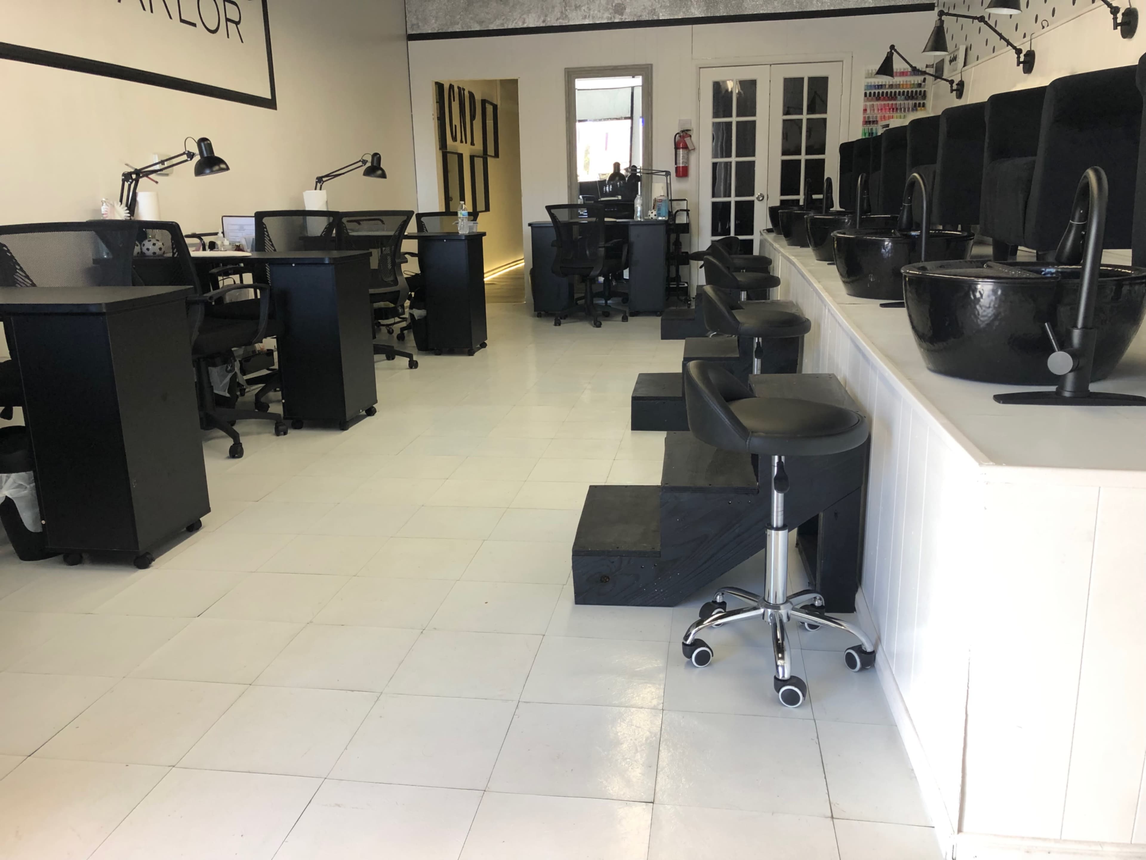 The image shows the interior of a nail salon with several workstations, black pedicure bowls, and modern chairs arranged in a spacious layout.