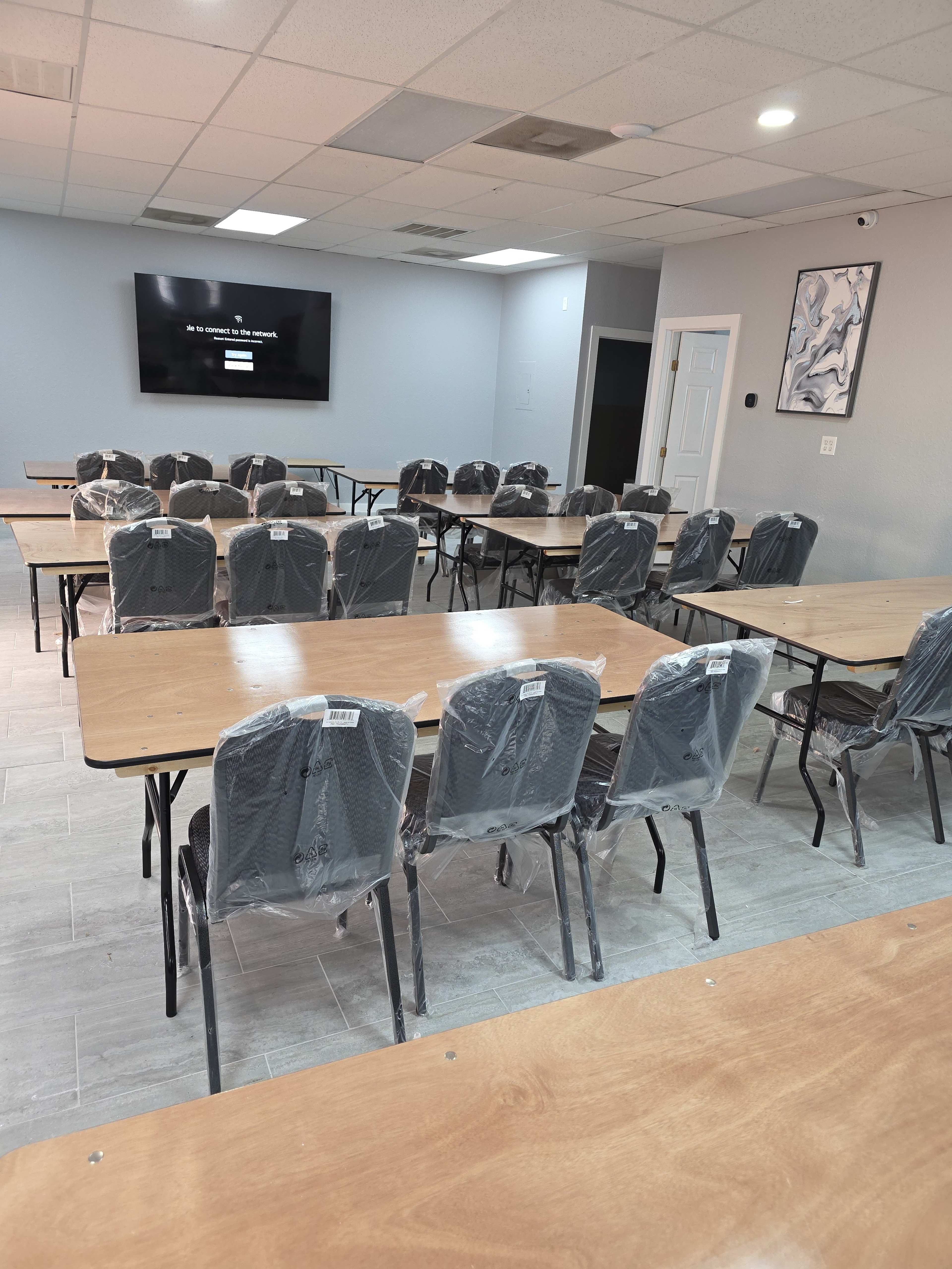 A meeting room with multiple tables and chairs, all covered in plastic wrap, is set up for an event, featuring a television screen mounted on the wall.