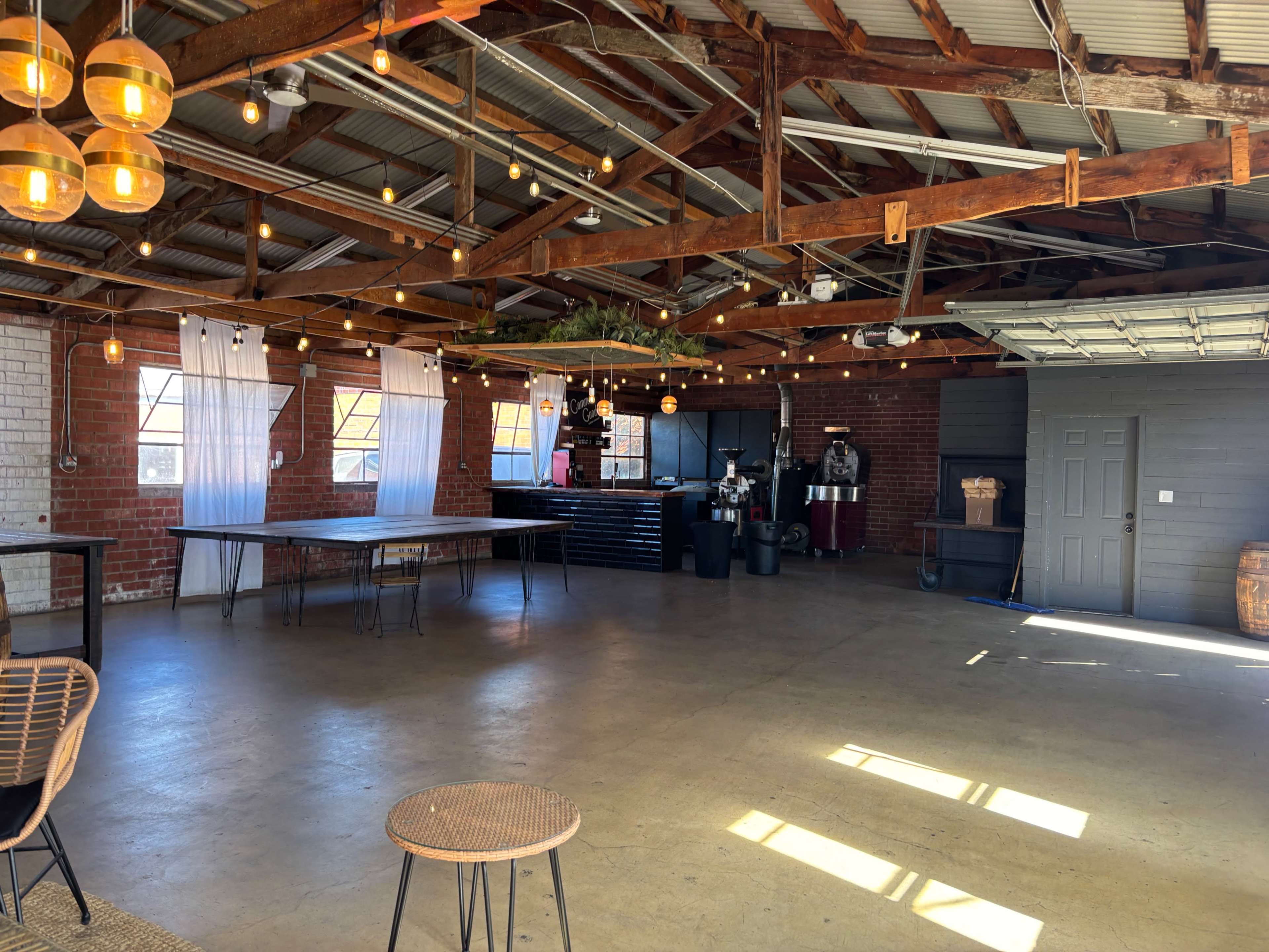 The image shows an empty rustic event space with exposed wooden beams, string lights, and a bar area, featuring large tables and a concrete floor.