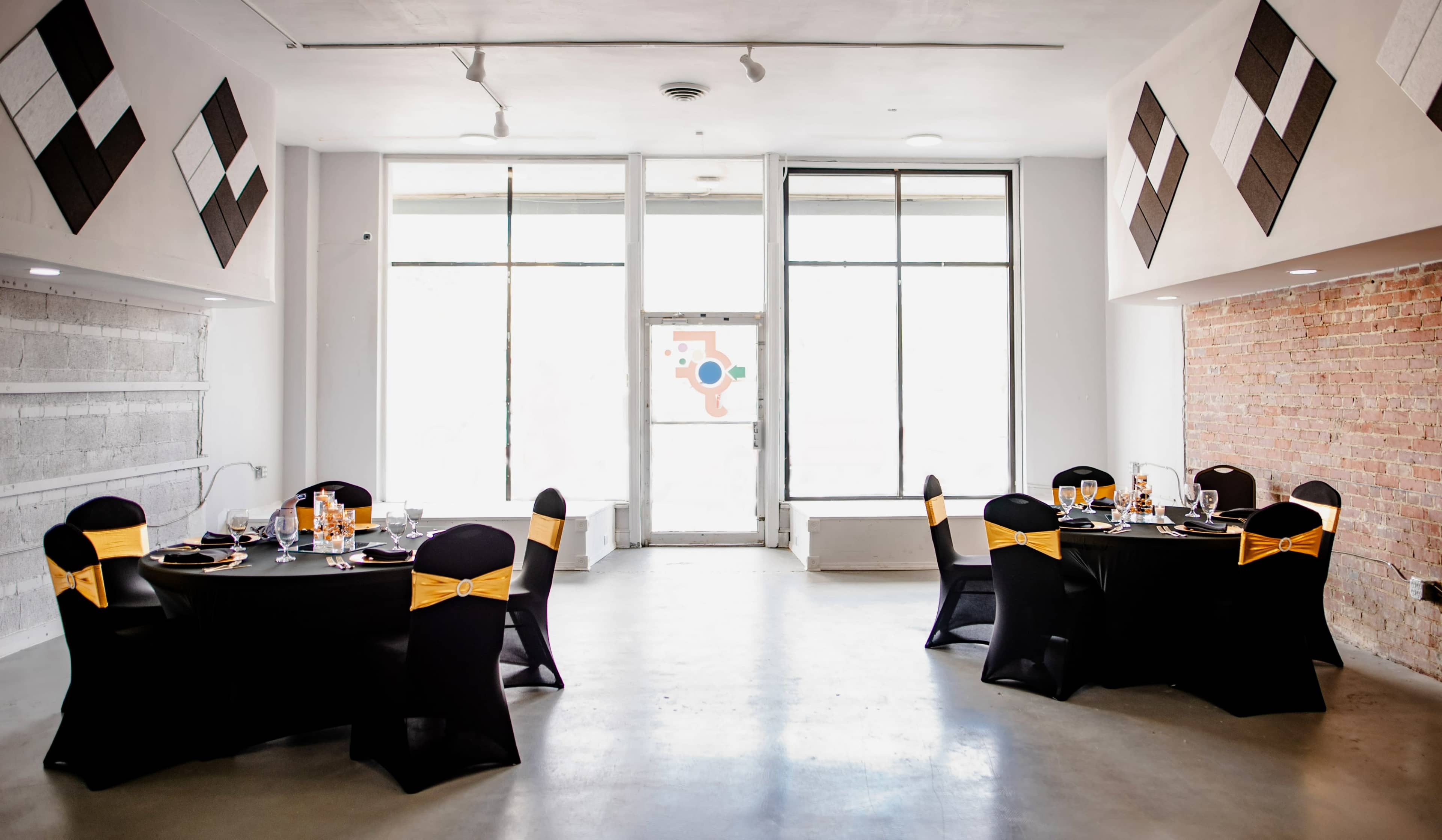 A modern event space with two round tables dressed in black tablecloths and yellow sashes, set against a backdrop of large windows and exposed brick walls.