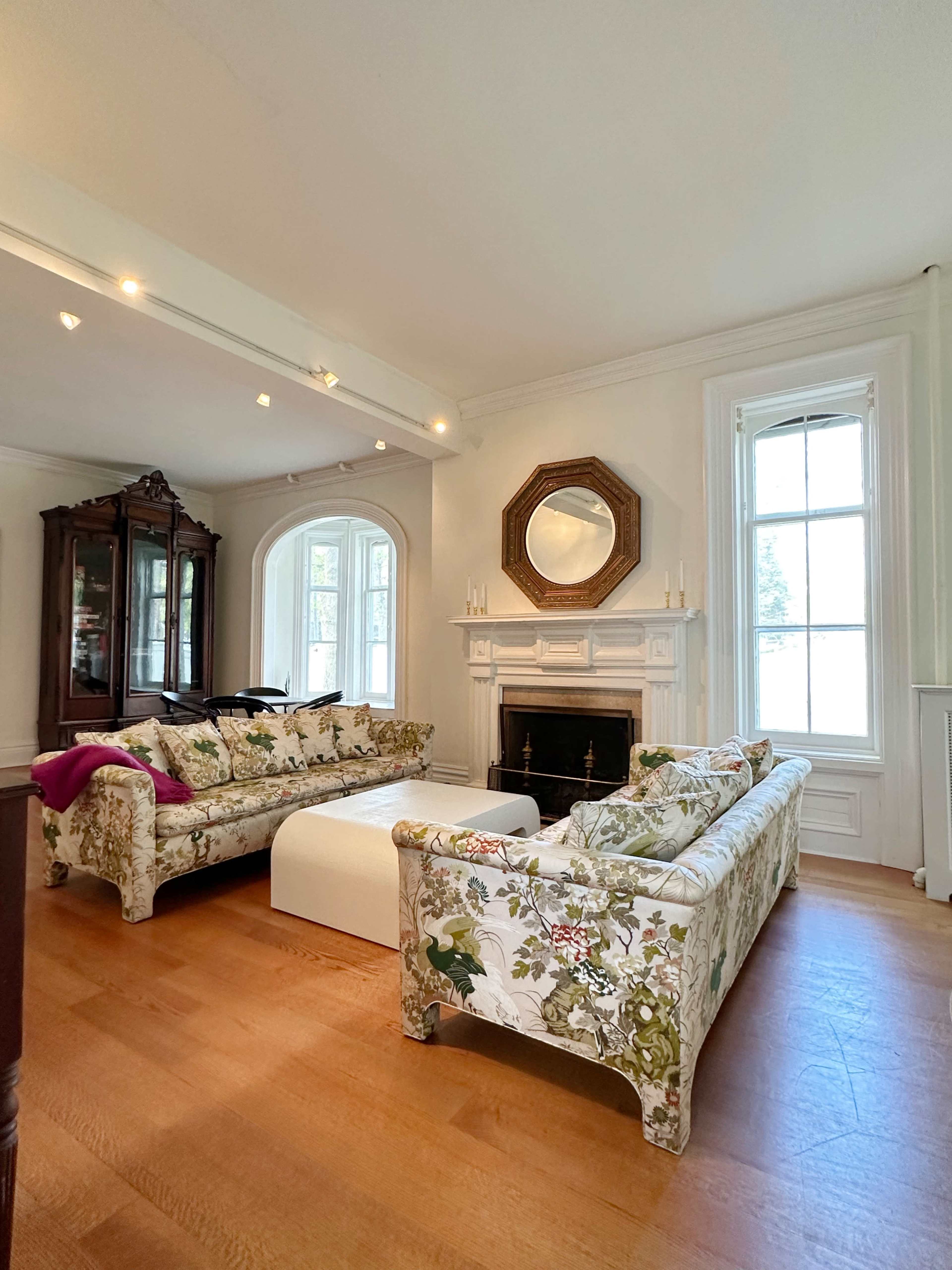 The living room features two floral-patterned sofas facing a white coffee table and a fireplace, with large windows letting in natural light.
