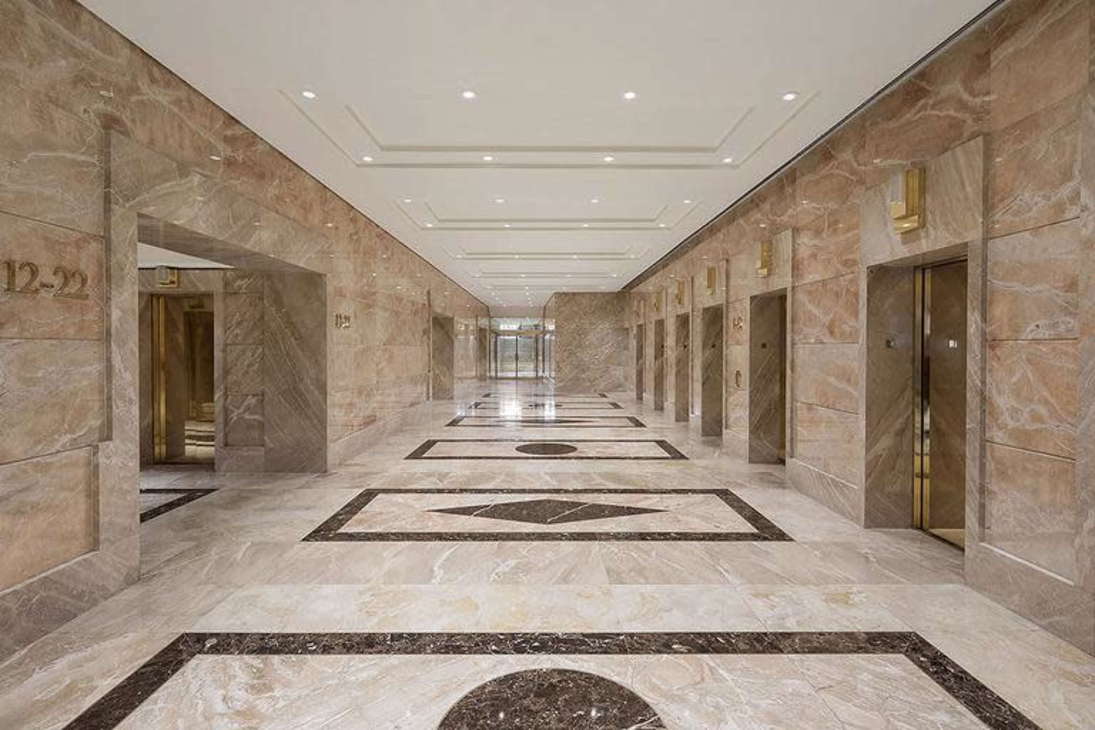 The image shows a wide hallway featuring multiple elevator doors lined on either side, with a marble floor displaying geometric patterns and subtle lighting above.