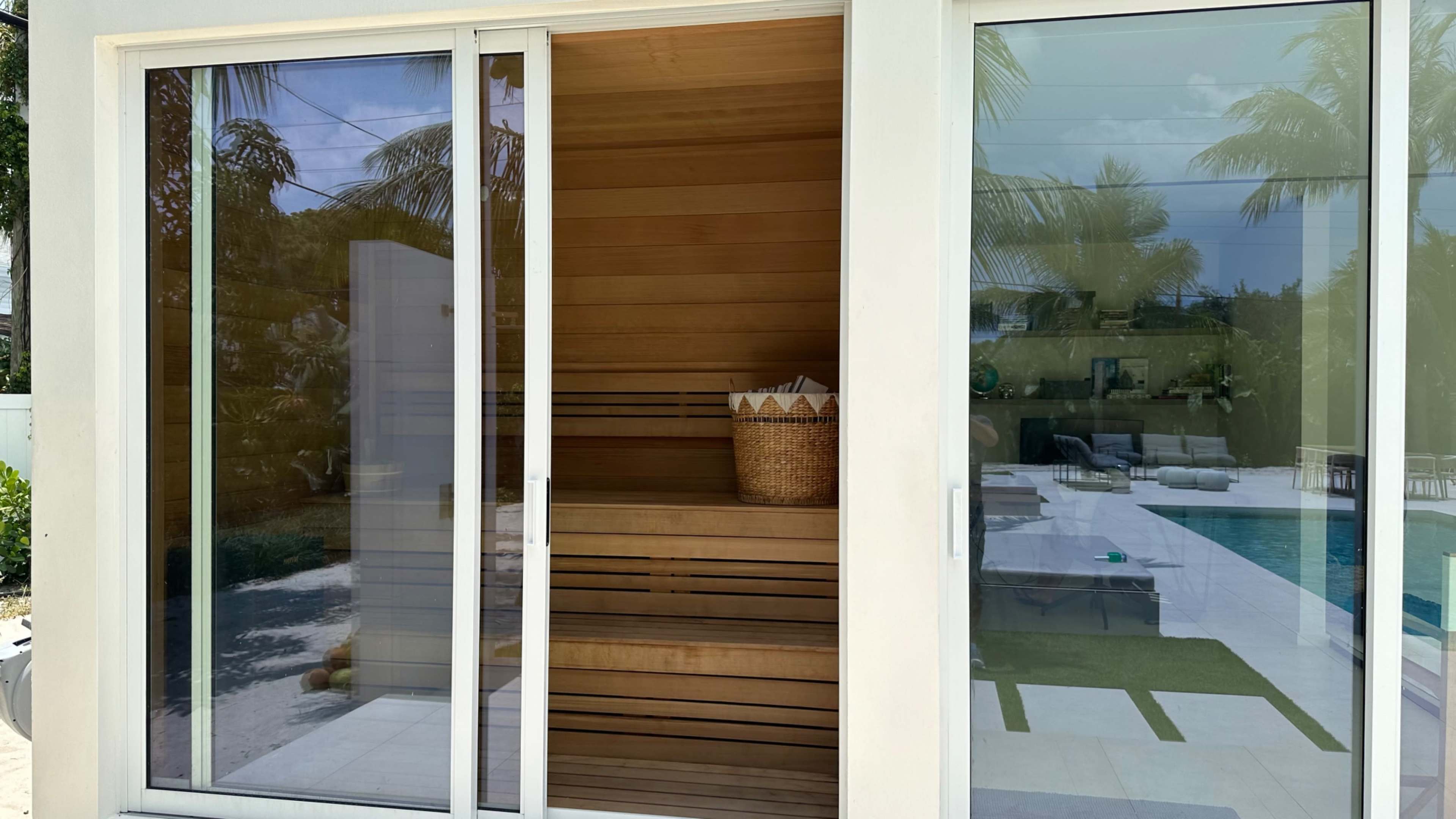 The image shows a modern sauna made of wood, located beside a large outdoor pool area surrounded by palm trees.