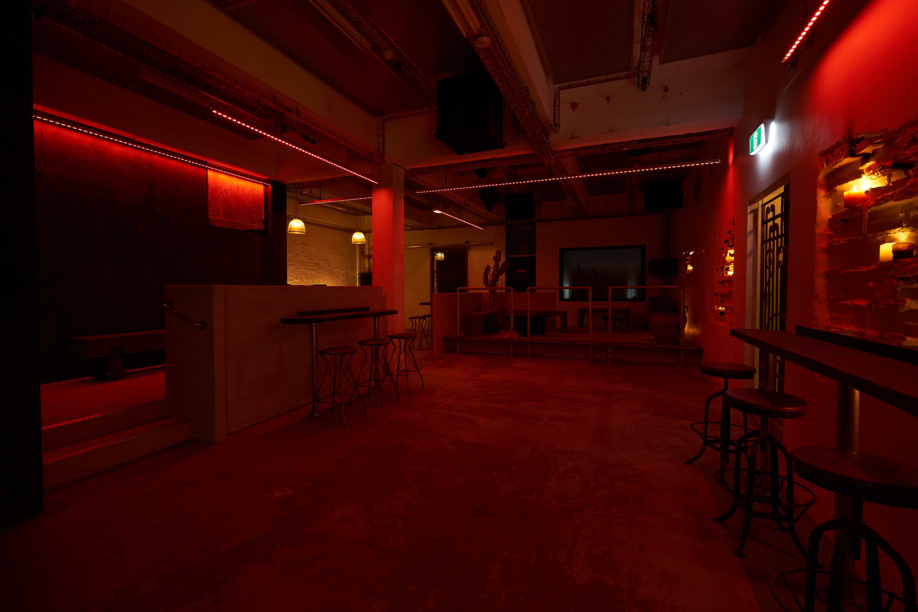 The image shows a dimly lit interior space with red lighting, featuring bar stools and a counter against textured walls.