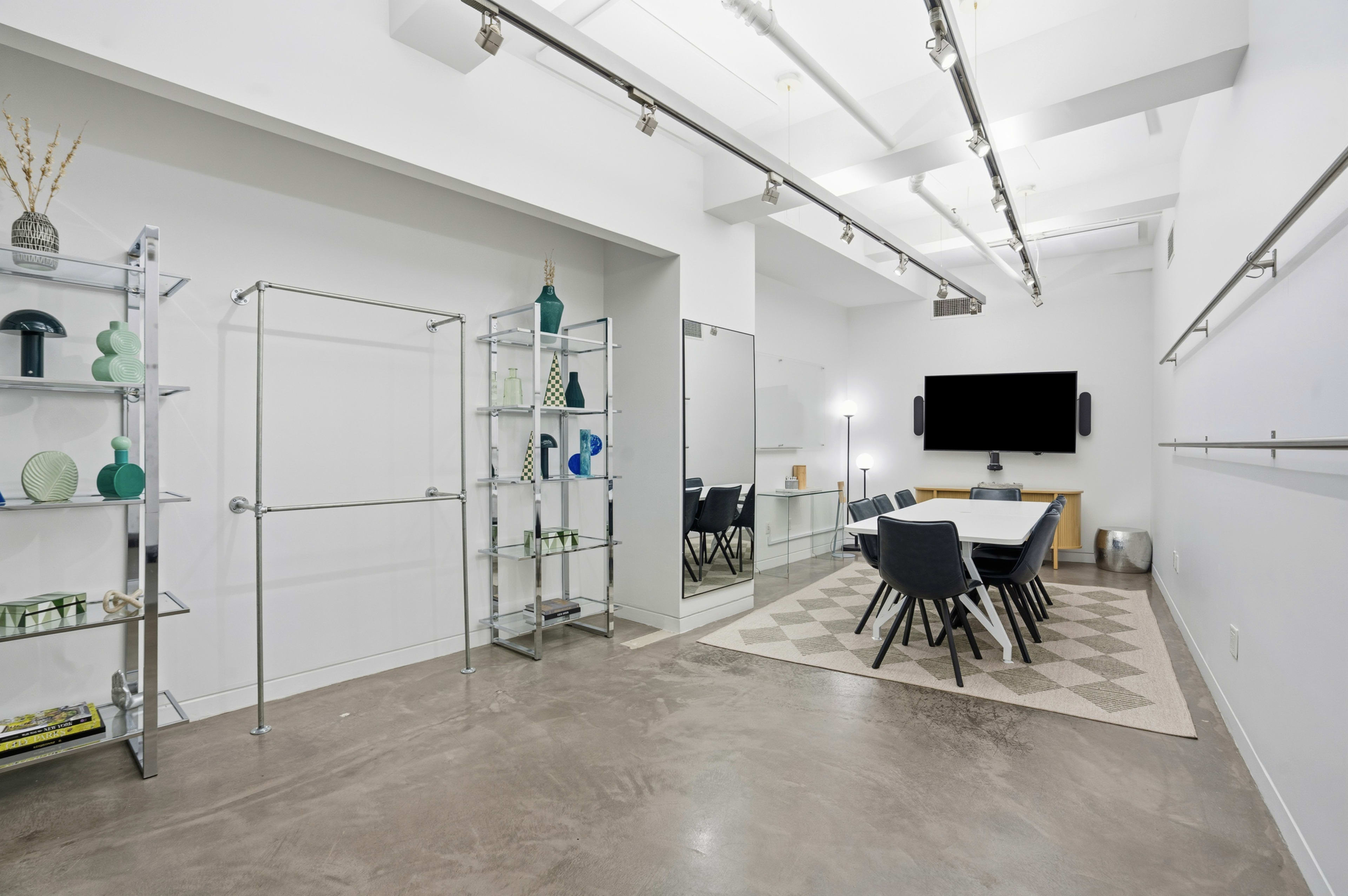 The image shows a modern meeting room with a rectangular table surrounded by chairs, a large television screen on the wall, and shelves displaying decorative items.