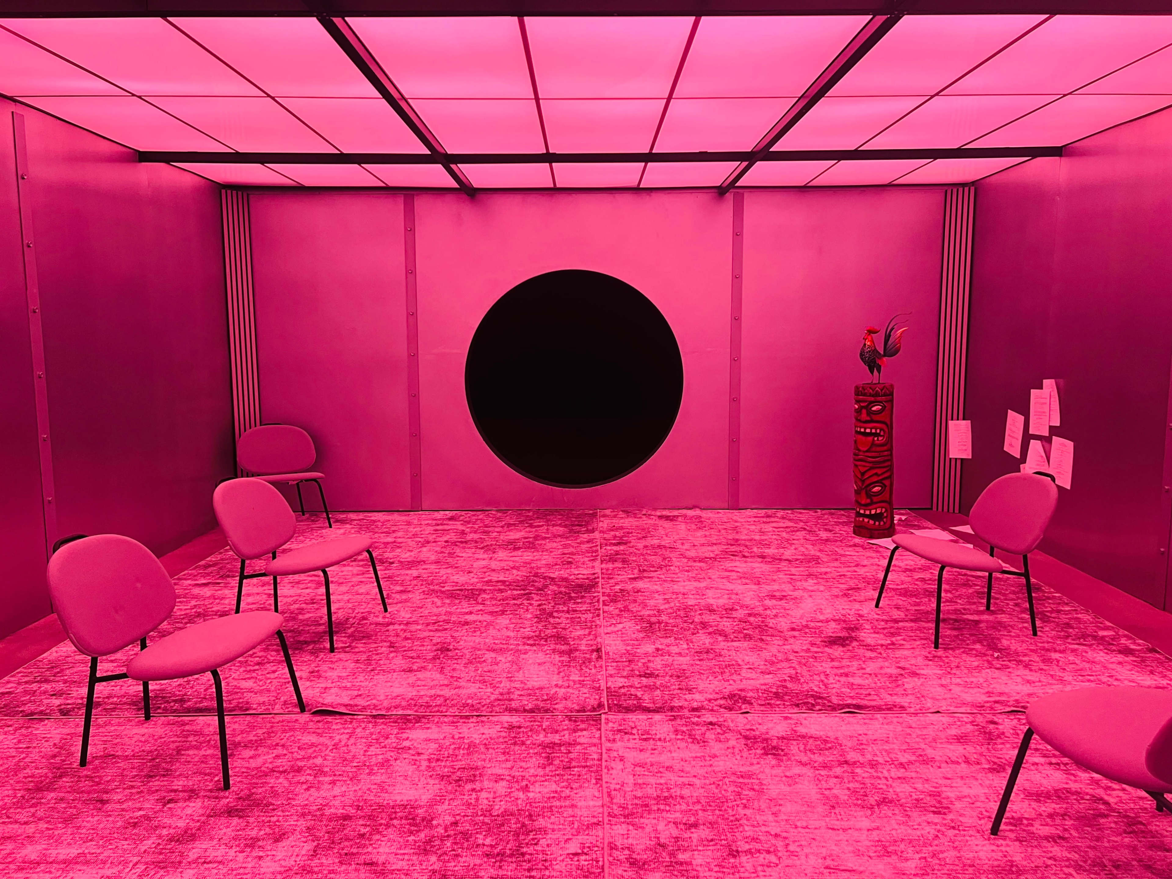The image shows a room illuminated in pink light with several chairs arranged around a circular opening in the wall.