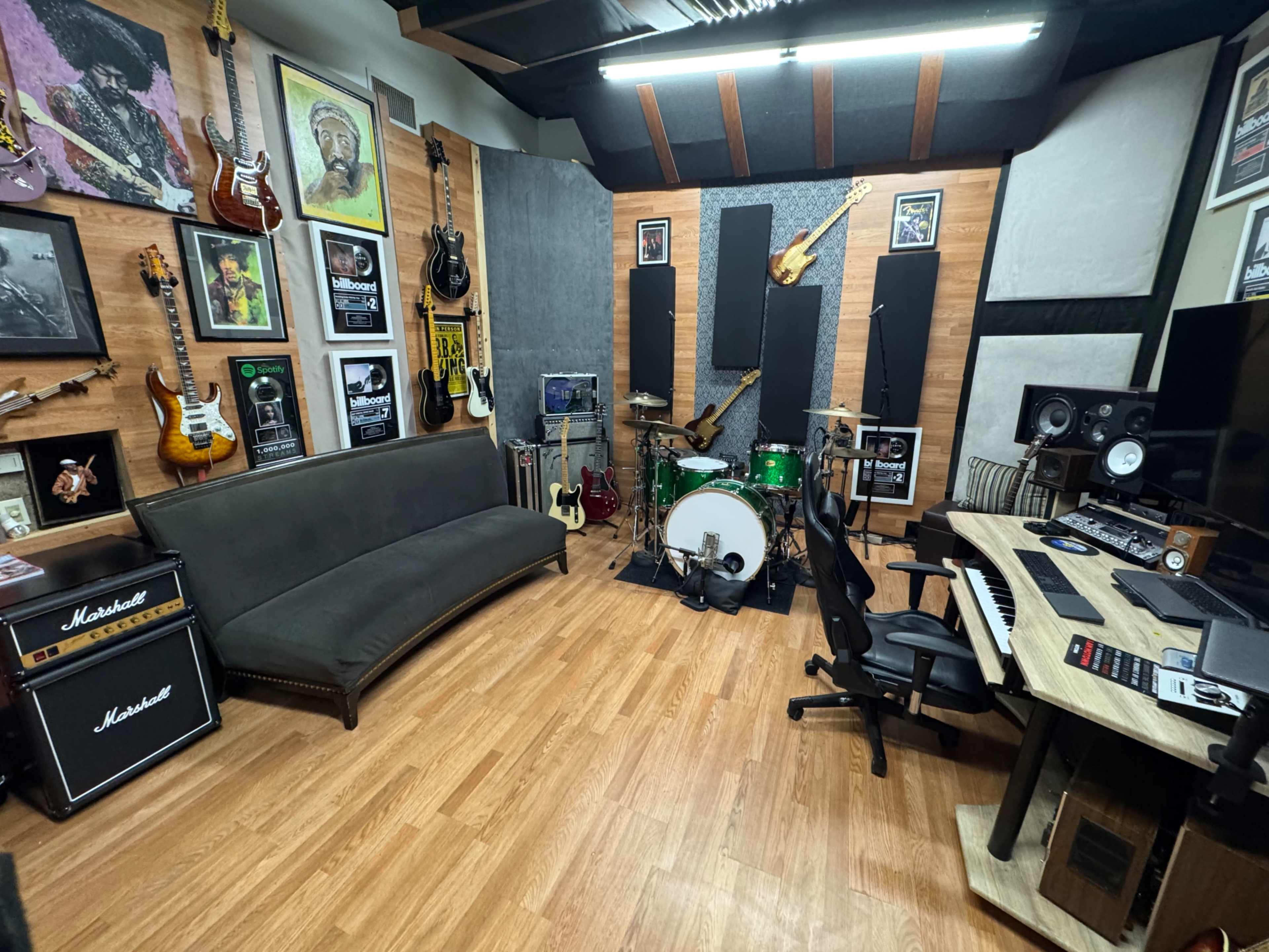 The image shows a music recording studio featuring guitars on the walls, a drum set, a keyboard, and a sofa, with soundproofing panels and artwork present.