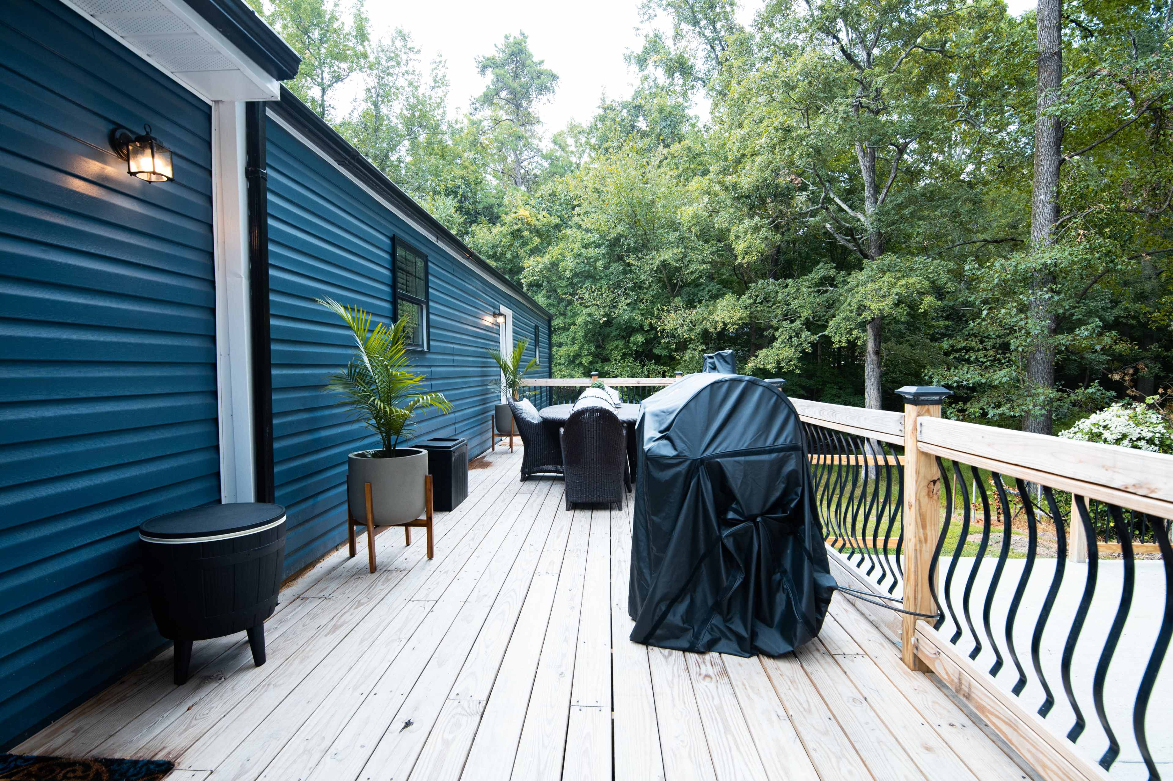 A wooden deck features a covered grill, outdoor seating, and potted plants, surrounded by trees.