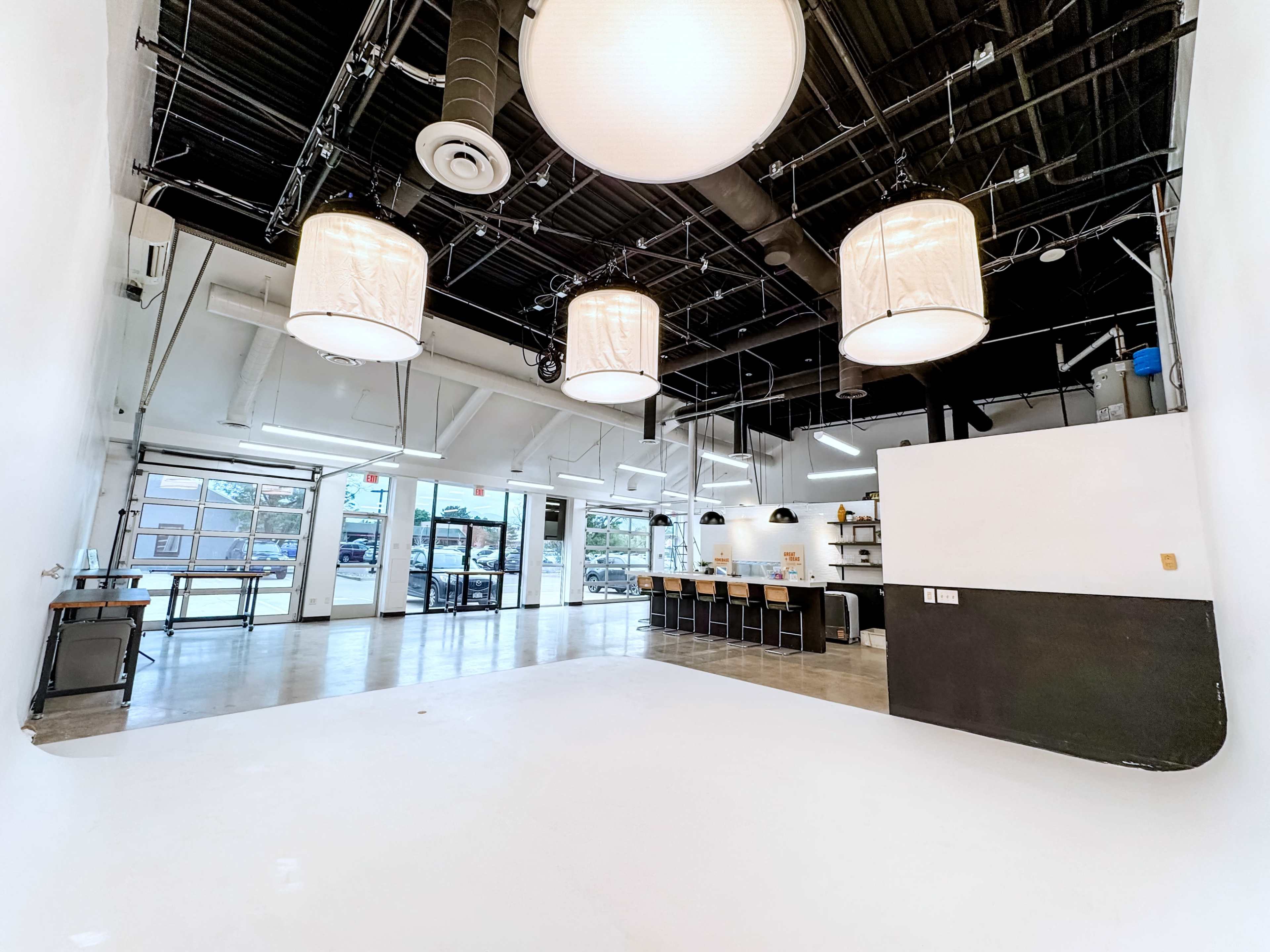 The image shows a spacious, well-lit interior featuring large pendant lights, a counter with seating, and large windows that provide natural light.