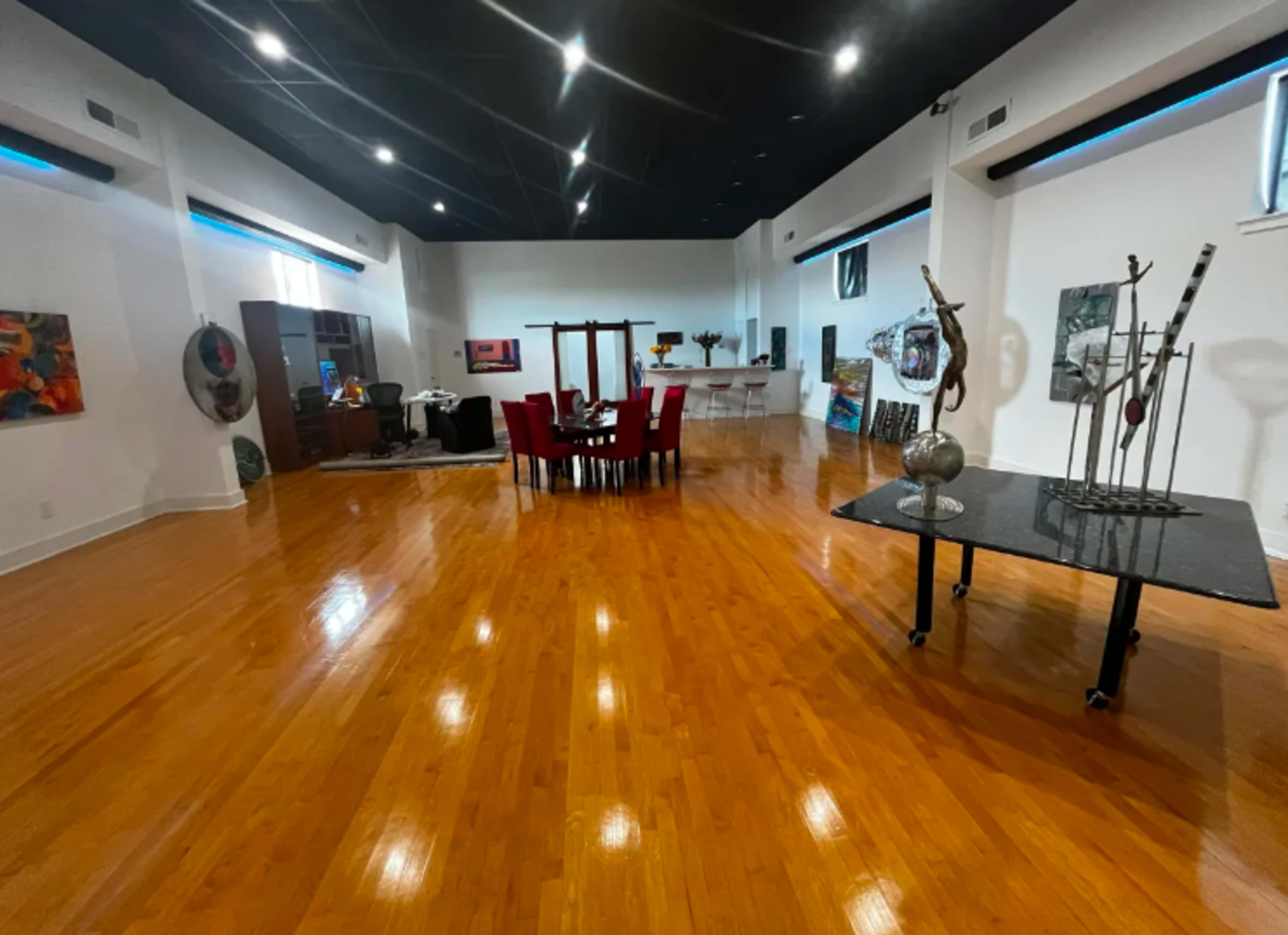 A spacious room with polished wooden floors, featuring a dining table surrounded by red chairs, an office area, and various artworks displayed on the walls.