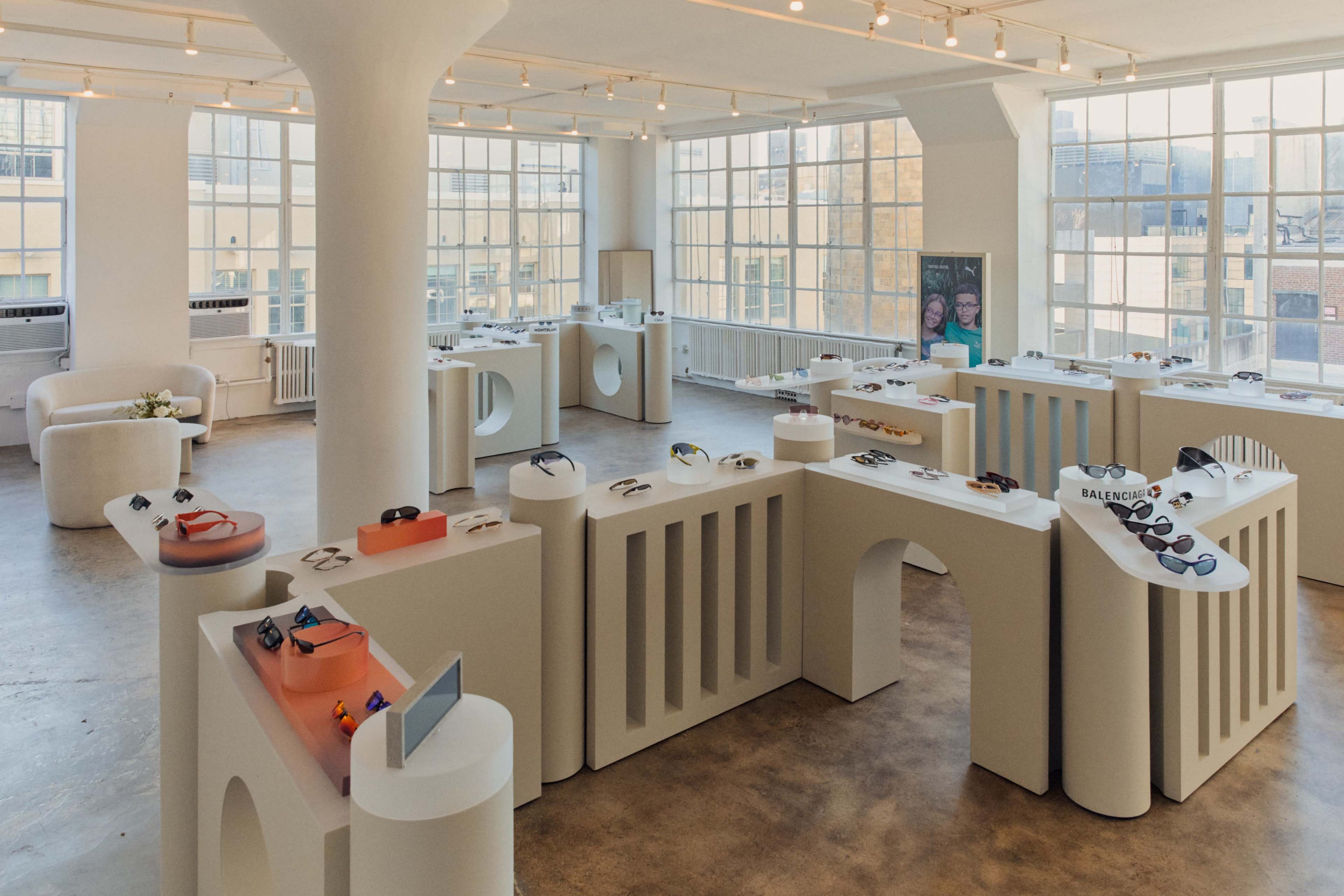 The image shows a modern, well-lit exhibition space with white display stands showcasing a variety of sunglasses.