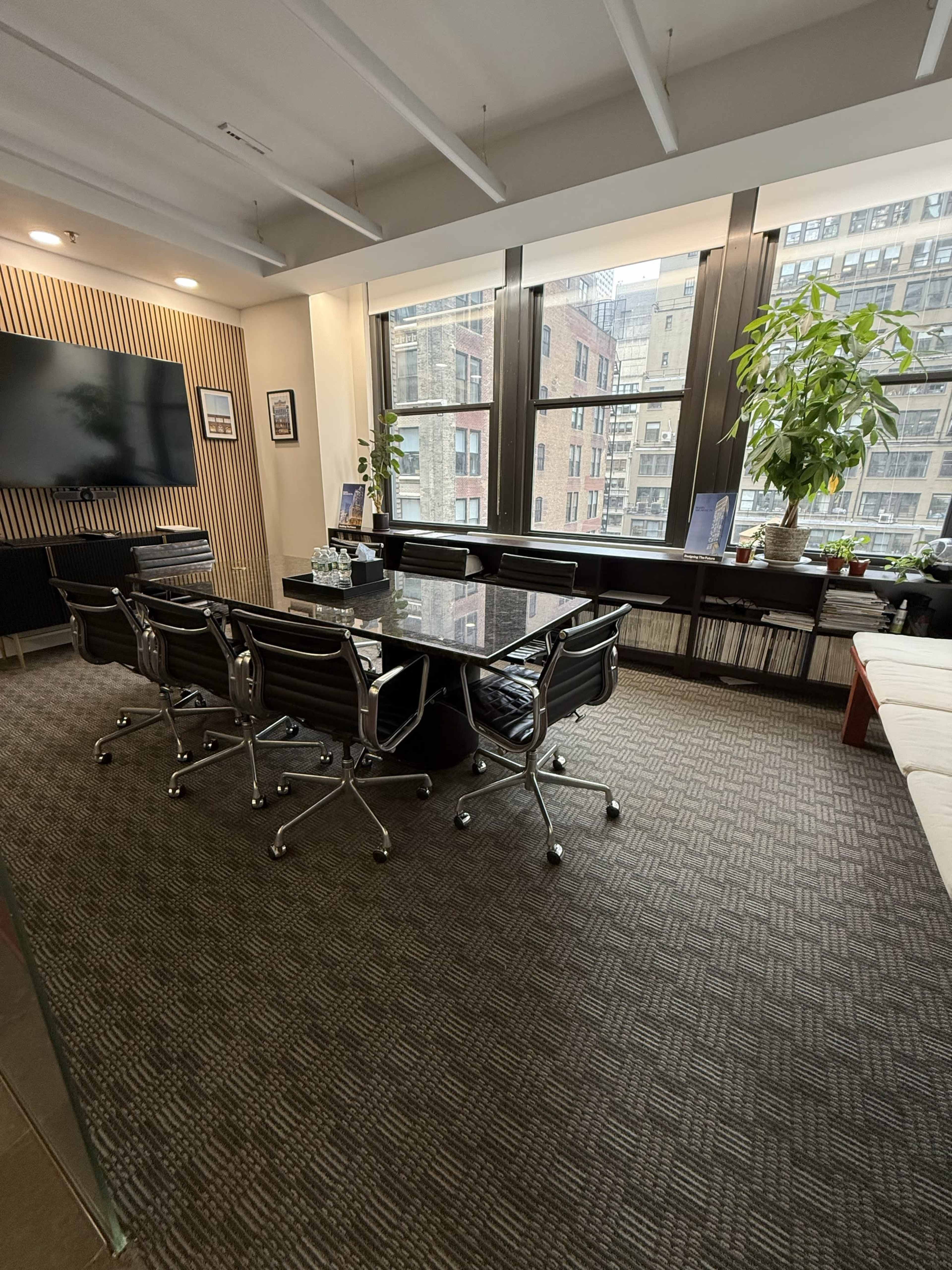 The image shows a modern office space with a conference table and several chairs, large windows allowing natural light, and decorative plants along the windowsill.