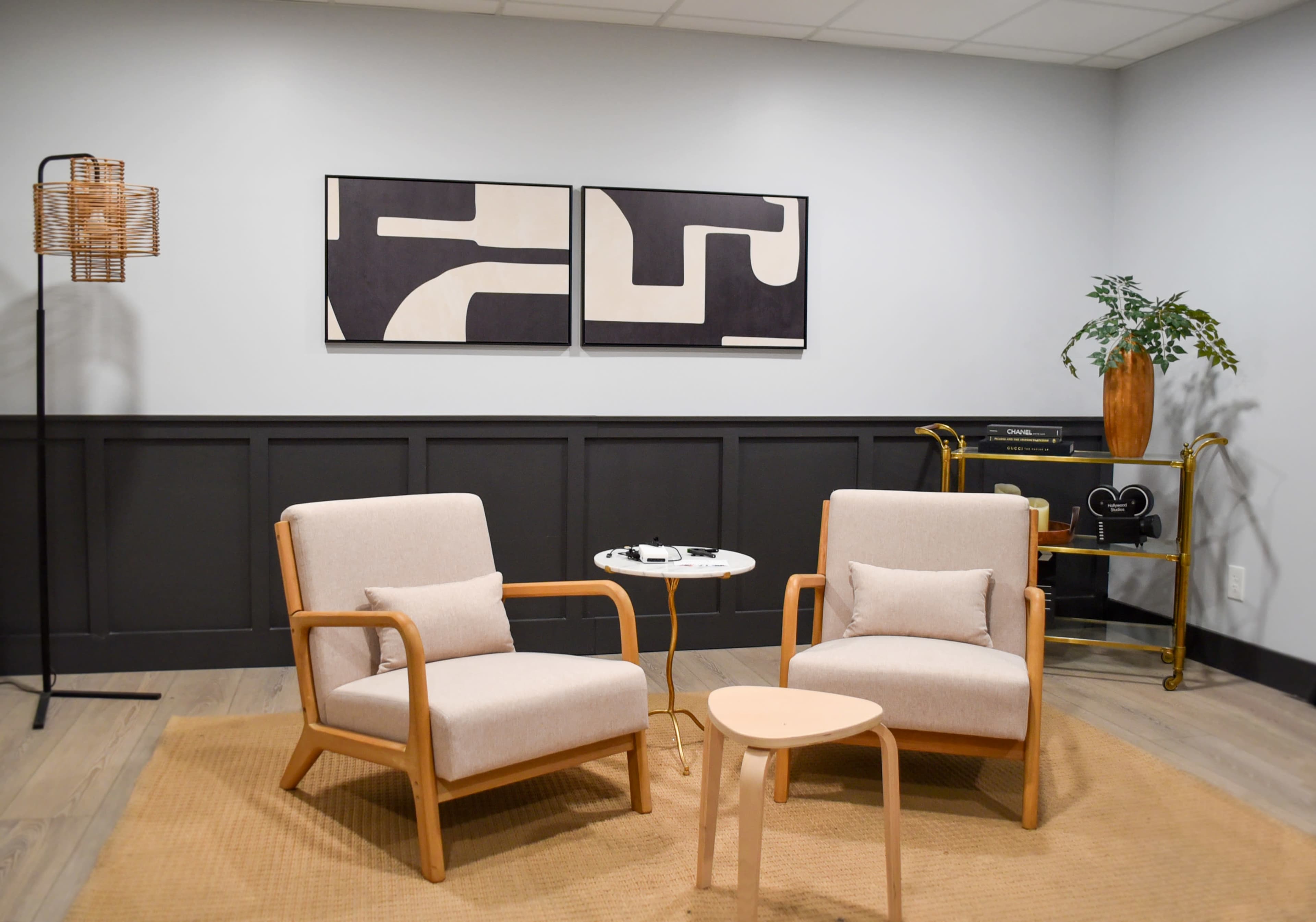The image shows a minimalist lounge area with two light-colored chairs, a small table, abstract artwork on the wall, and a decorative plant.