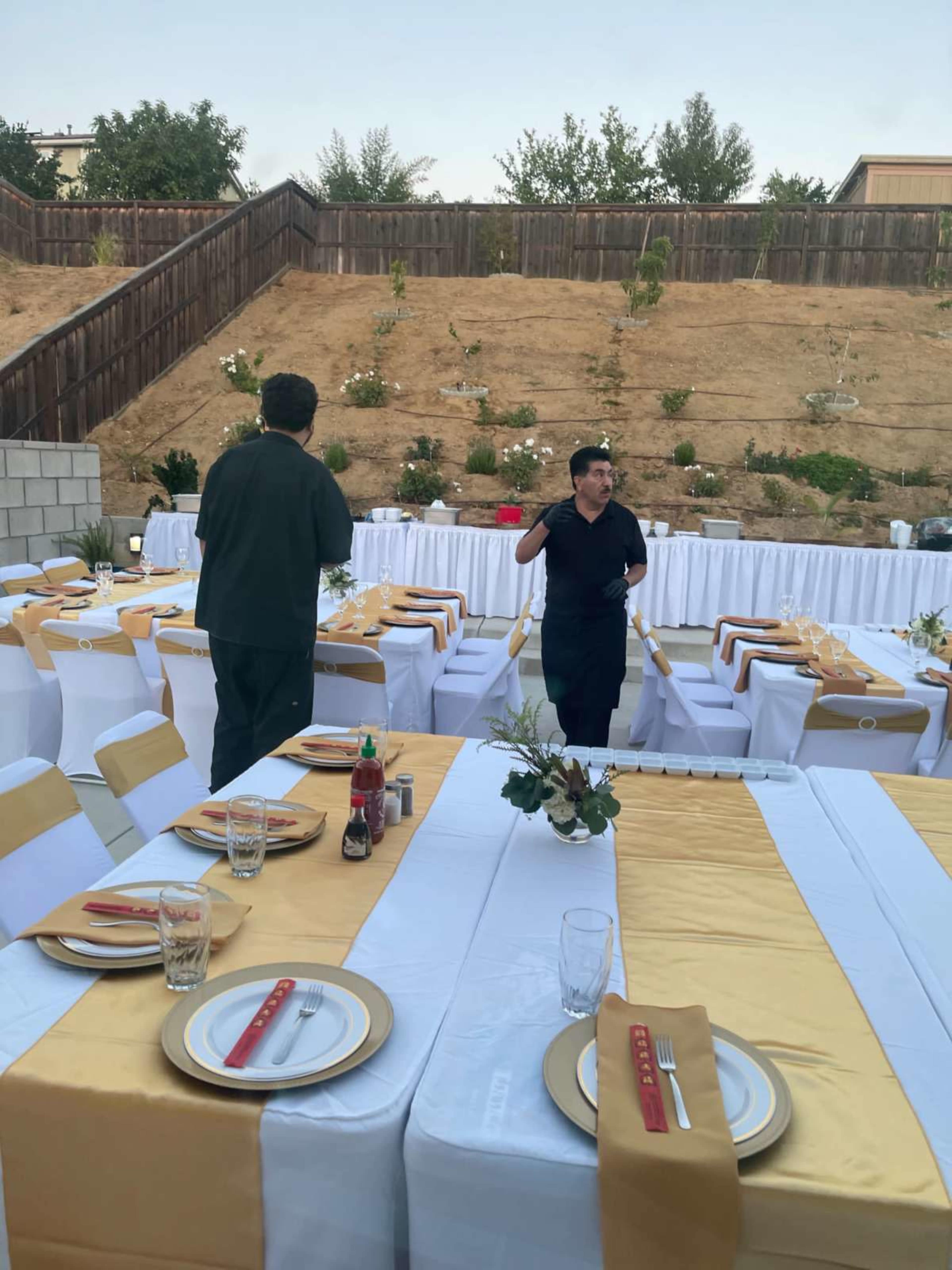 Two people set up a dining area with tables covered in white and gold tablecloths in a backyard.
