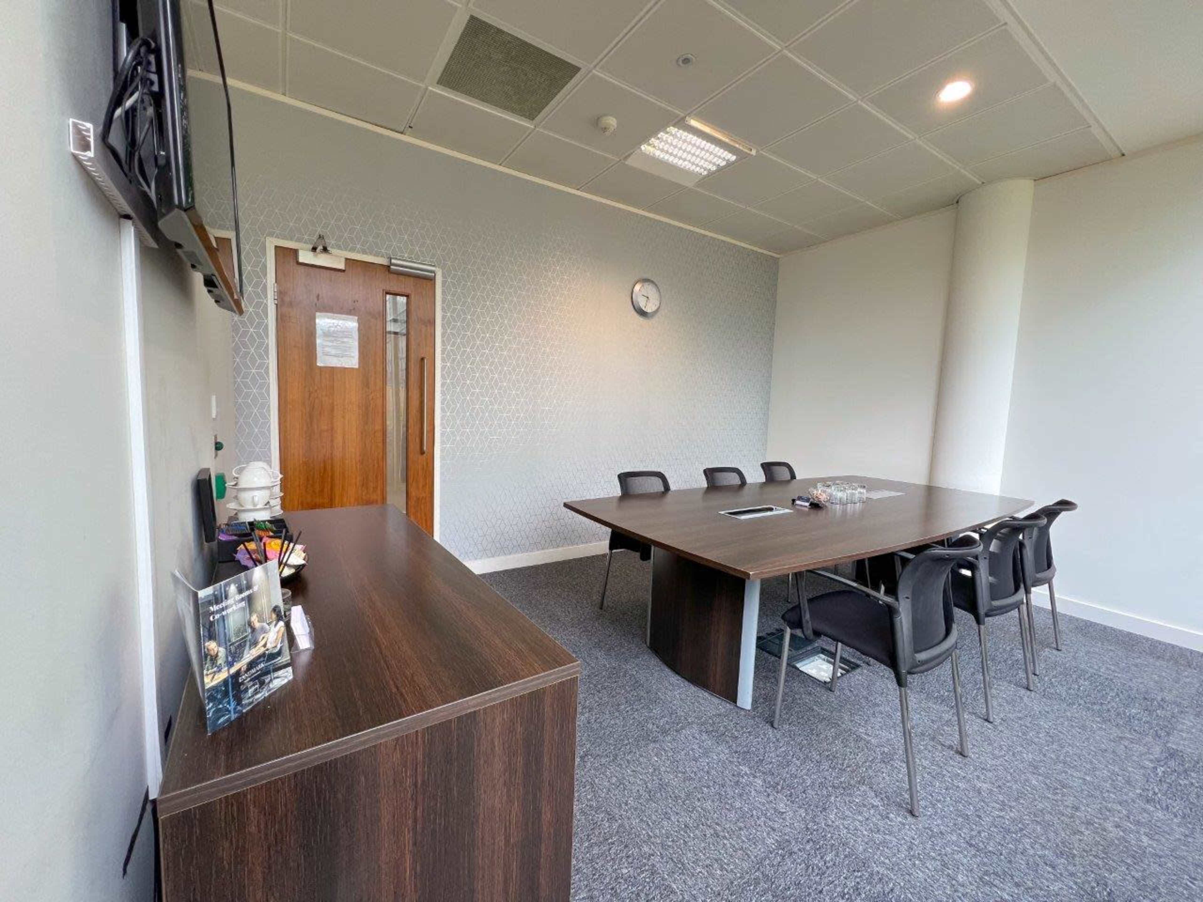The image shows a meeting room with a large table surrounded by black chairs, a wooden side cabinet, and a wall-mounted television.