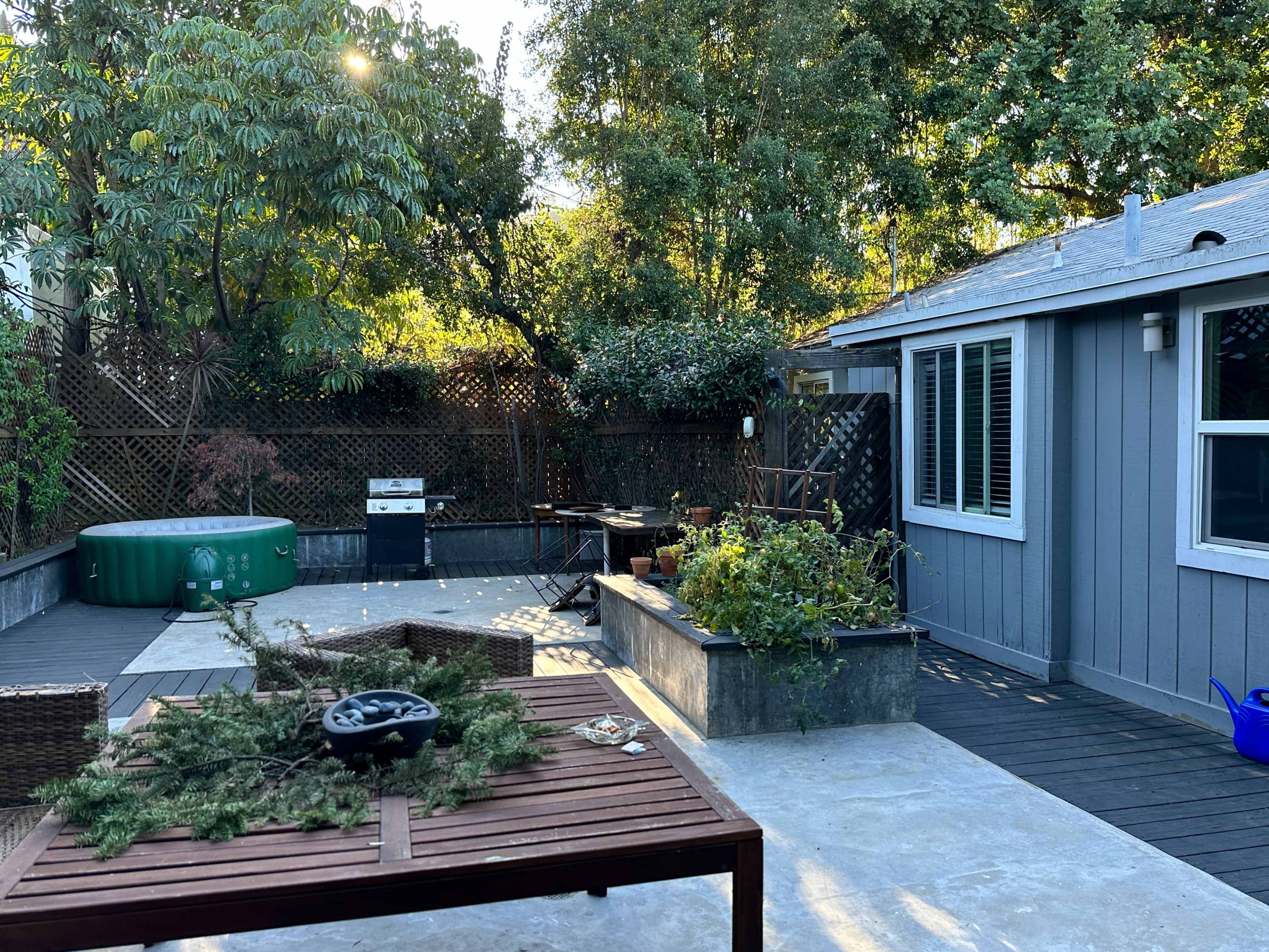 The image shows a backyard with a wooden patio, a green hot tub, a grill, and a seating area surrounded by trees and plants.