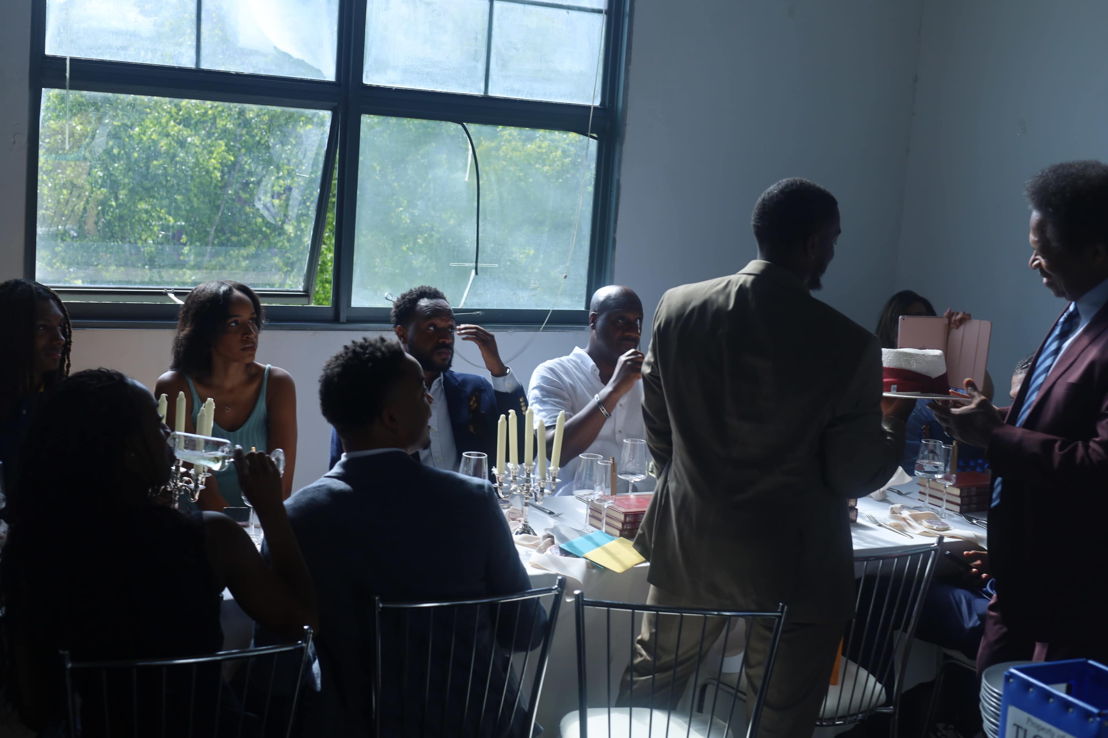 A group of people sit at a long table in a brightly lit room, engaged in conversation while some are enjoying food and drinks.