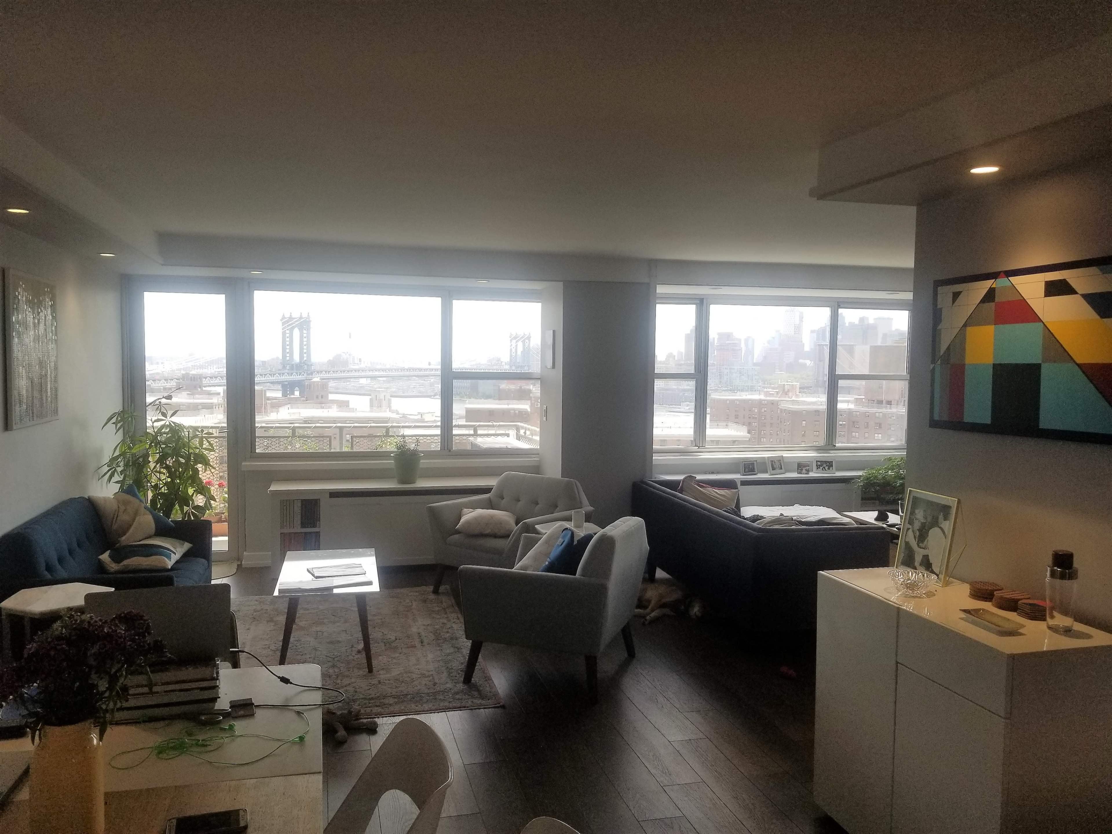 A modern living room features two sofas, a coffee table, and large windows showcasing a view of the Manhattan skyline and the Brooklyn Bridge.
