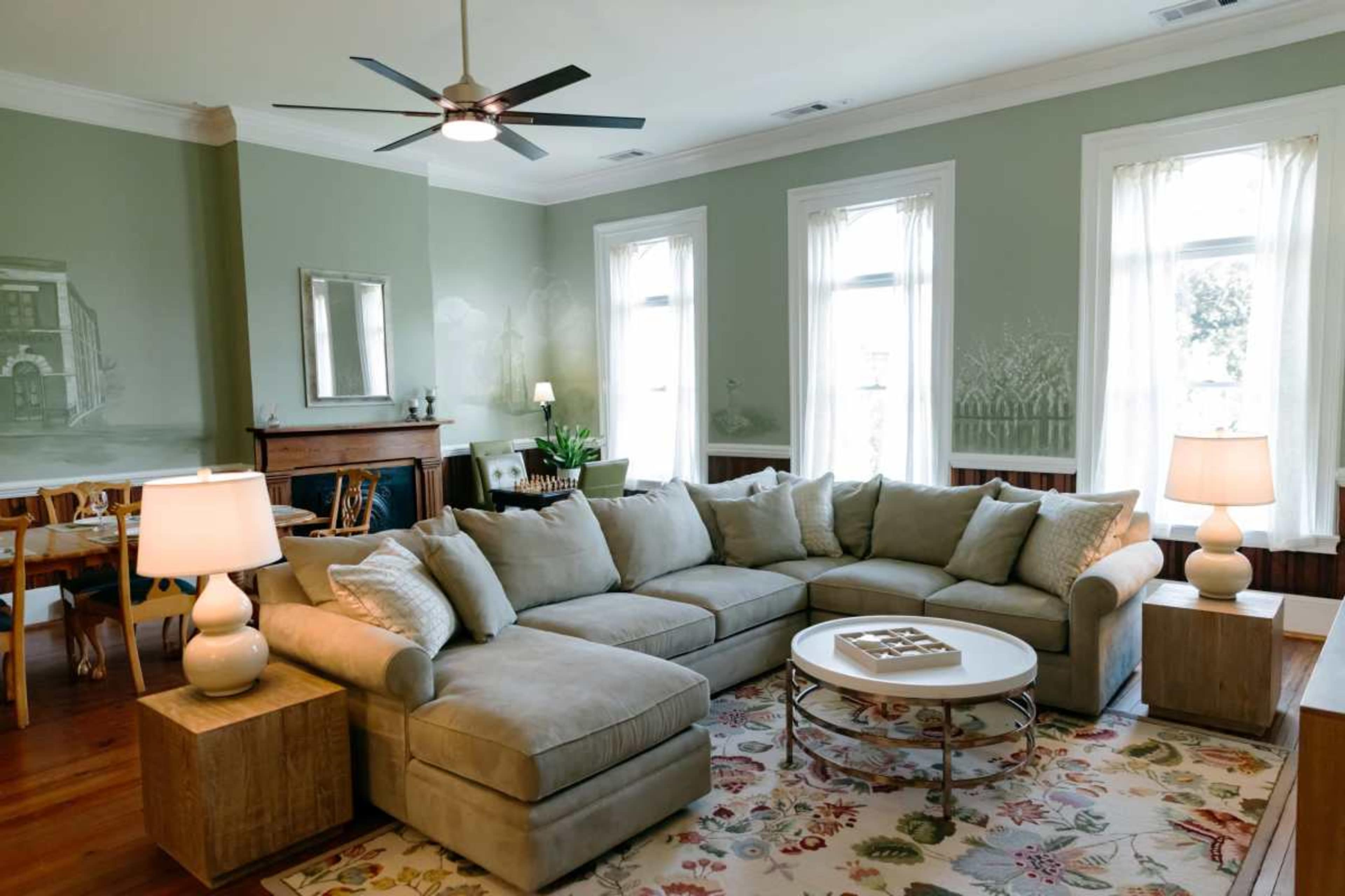 The living room features a large sectional sofa, a round coffee table, and two lamps, with beige walls and large windows allowing natural light to enter.