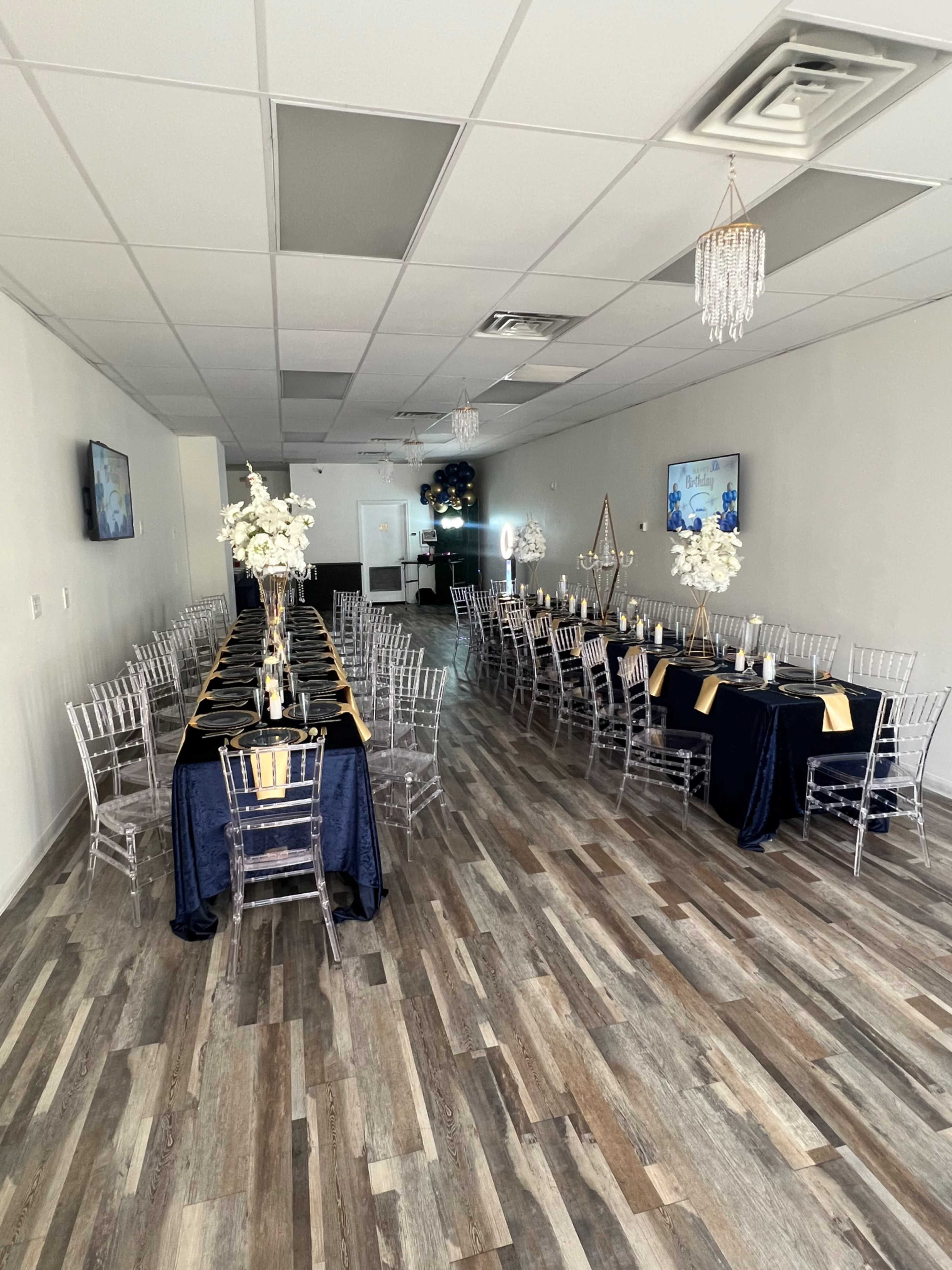 The image shows a banquet hall arranged with long tables covered in dark blue tablecloths, clear acrylic chairs, and centerpieces featuring white flowers.
