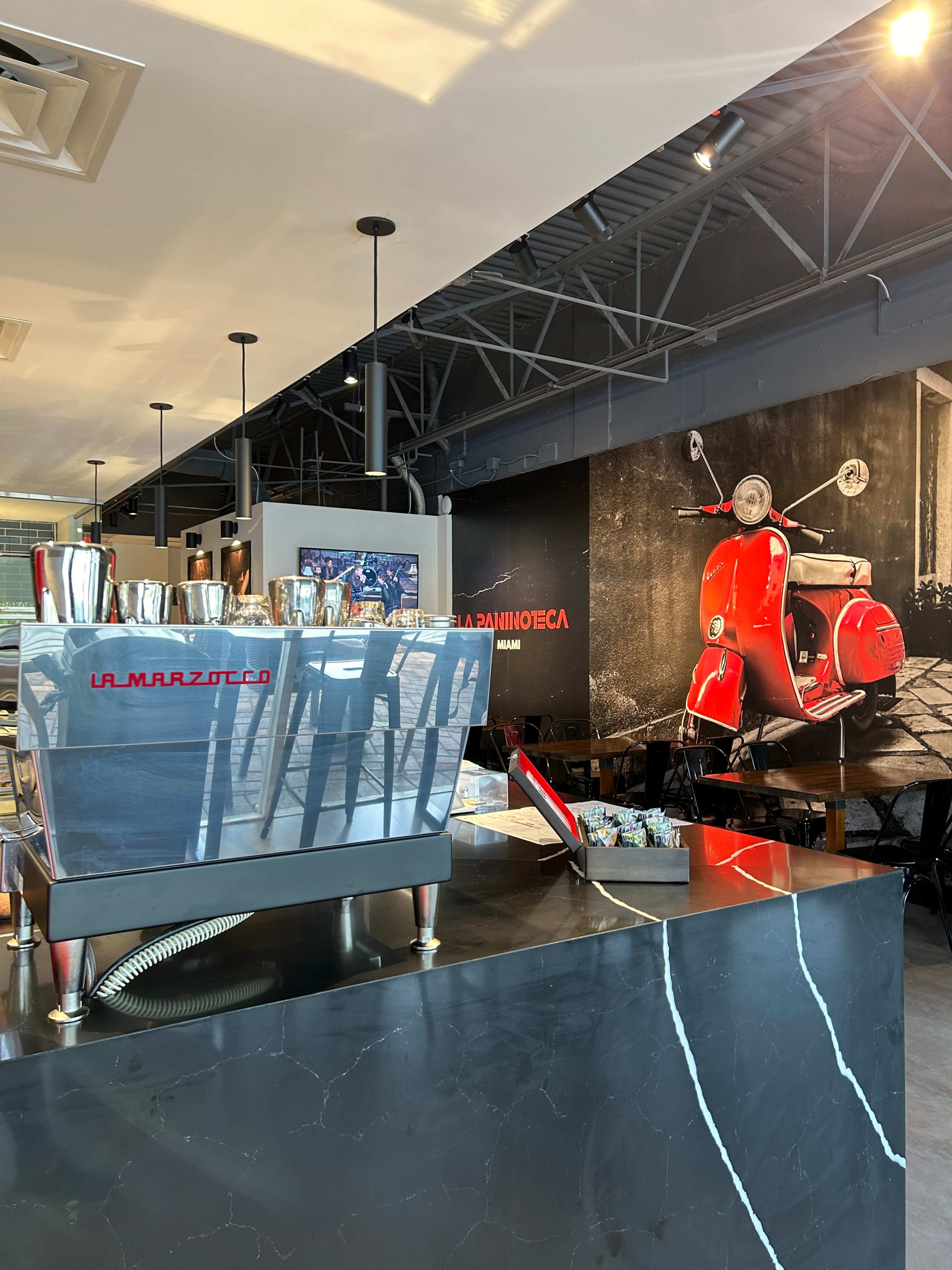 The image shows the interior of a café featuring a sleek coffee machine on a marble countertop, with a large mural of a red scooter on the wall.